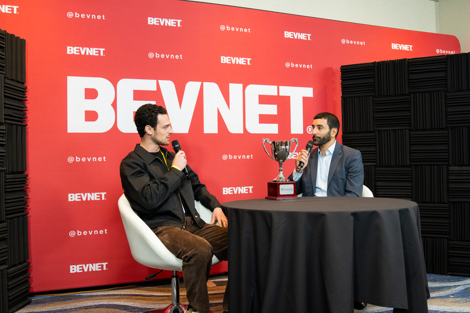 Two men seated at a small round table in front of a red BevNET backdrop with a silver trophy on the table.
