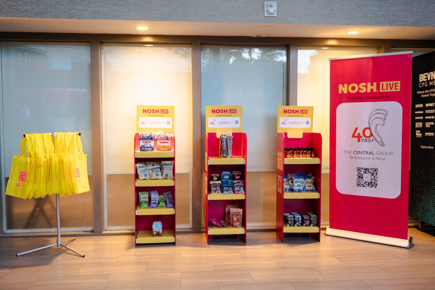 Three red and yellow product racks next to a Nosh Live banner for The Central Group and a rack of yellow tote bags.