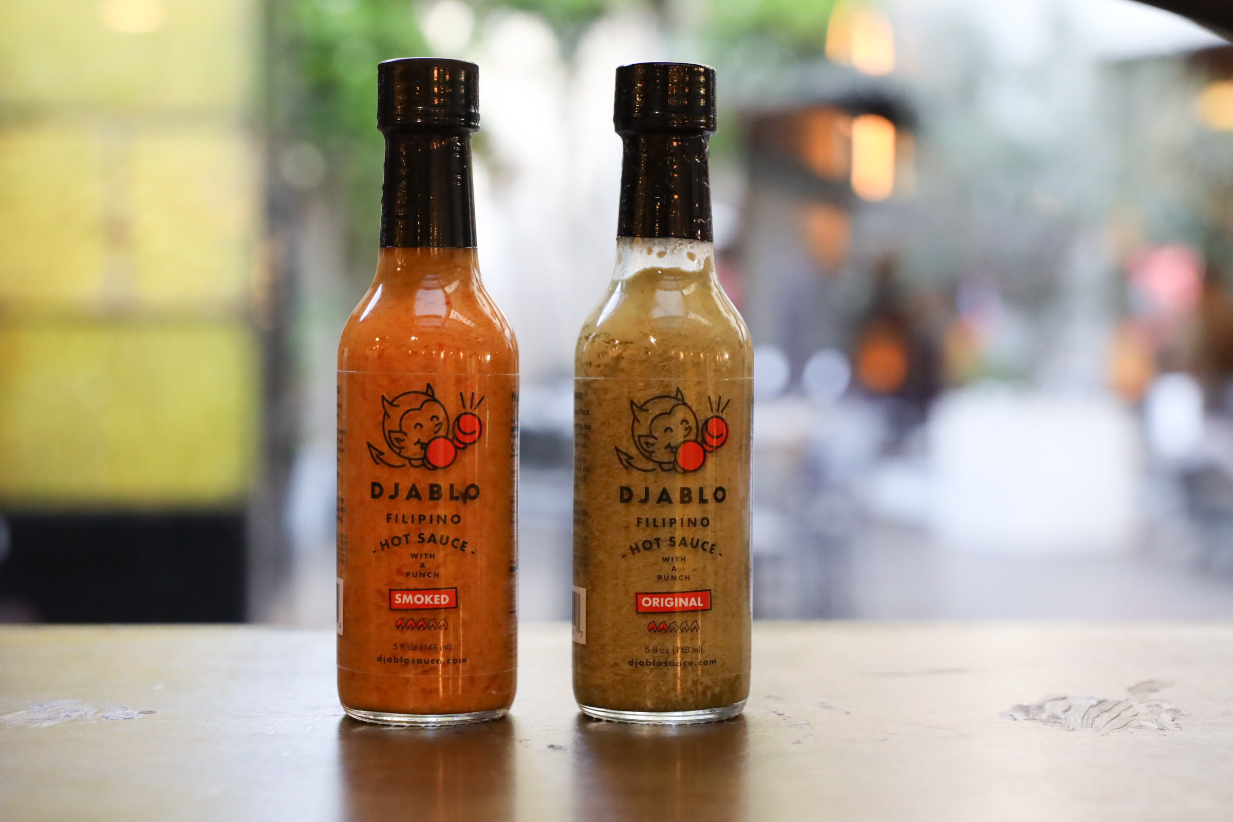 Two bottles of Djablo hot sauce, one orange and one tan, on a wooden surface.