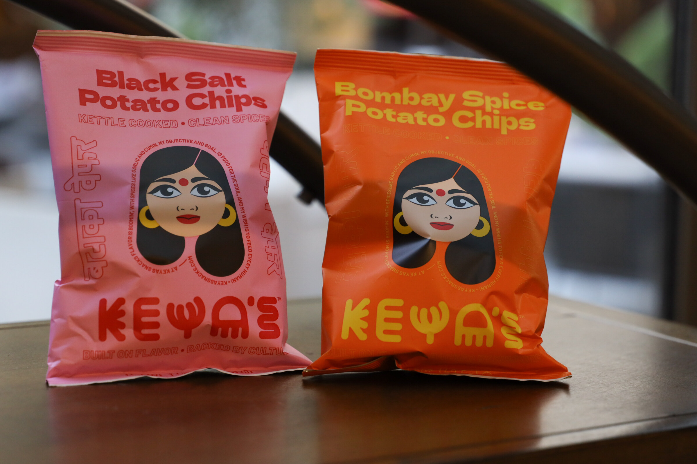Two bags of Keya's potato chips, one pink and one orange, on a wooden table.