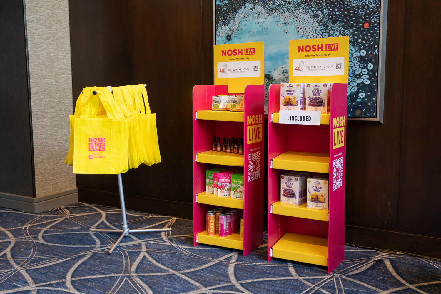 Two pink and yellow sampling racks holding snacks and bottles beside a rack of yellow tote bags.
