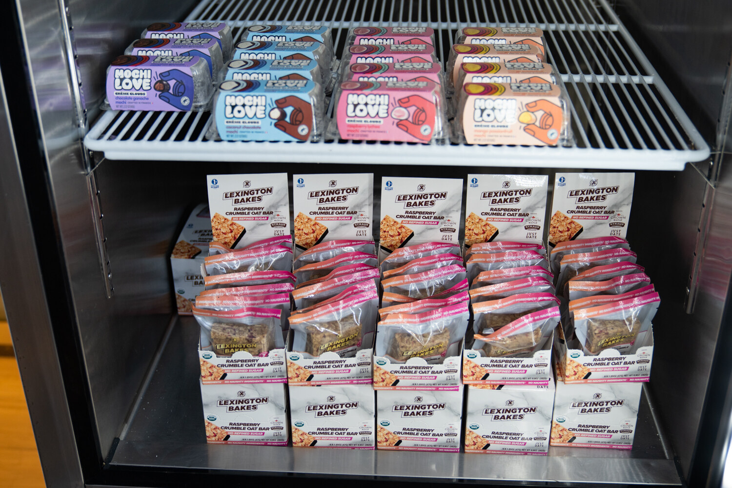 A glass-door refrigerator shelf with Mochi Love packs above boxes of Lexington Bakes crumble bars.