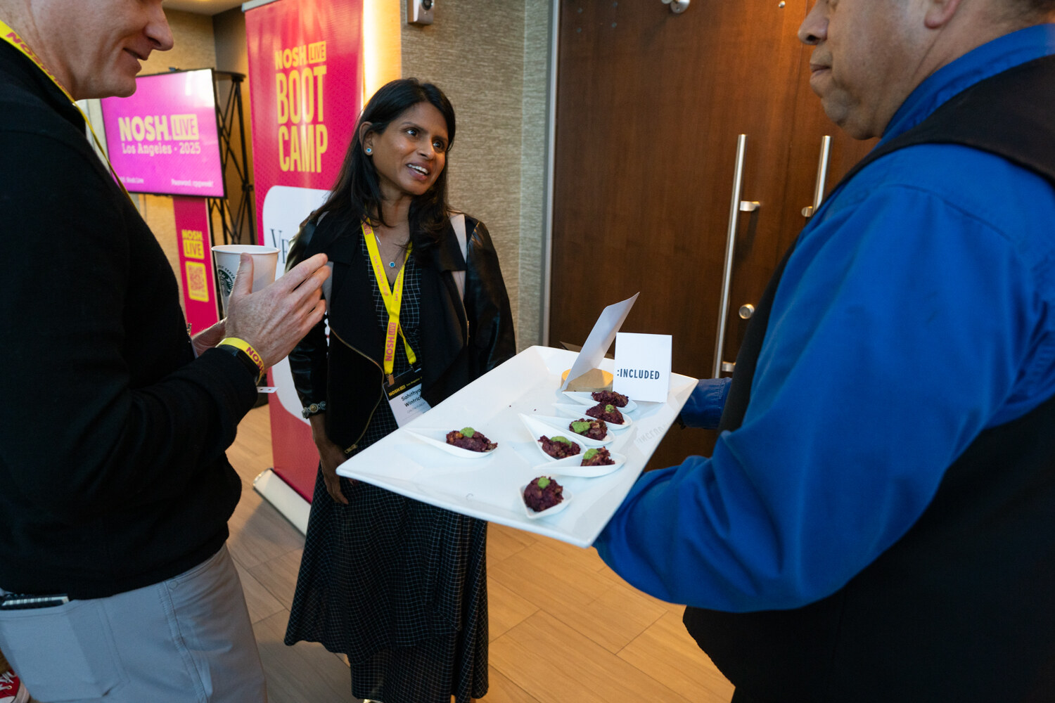 A server in a blue jacket holding a tray of chocolate bites while an attendee in a black jacket looks on.