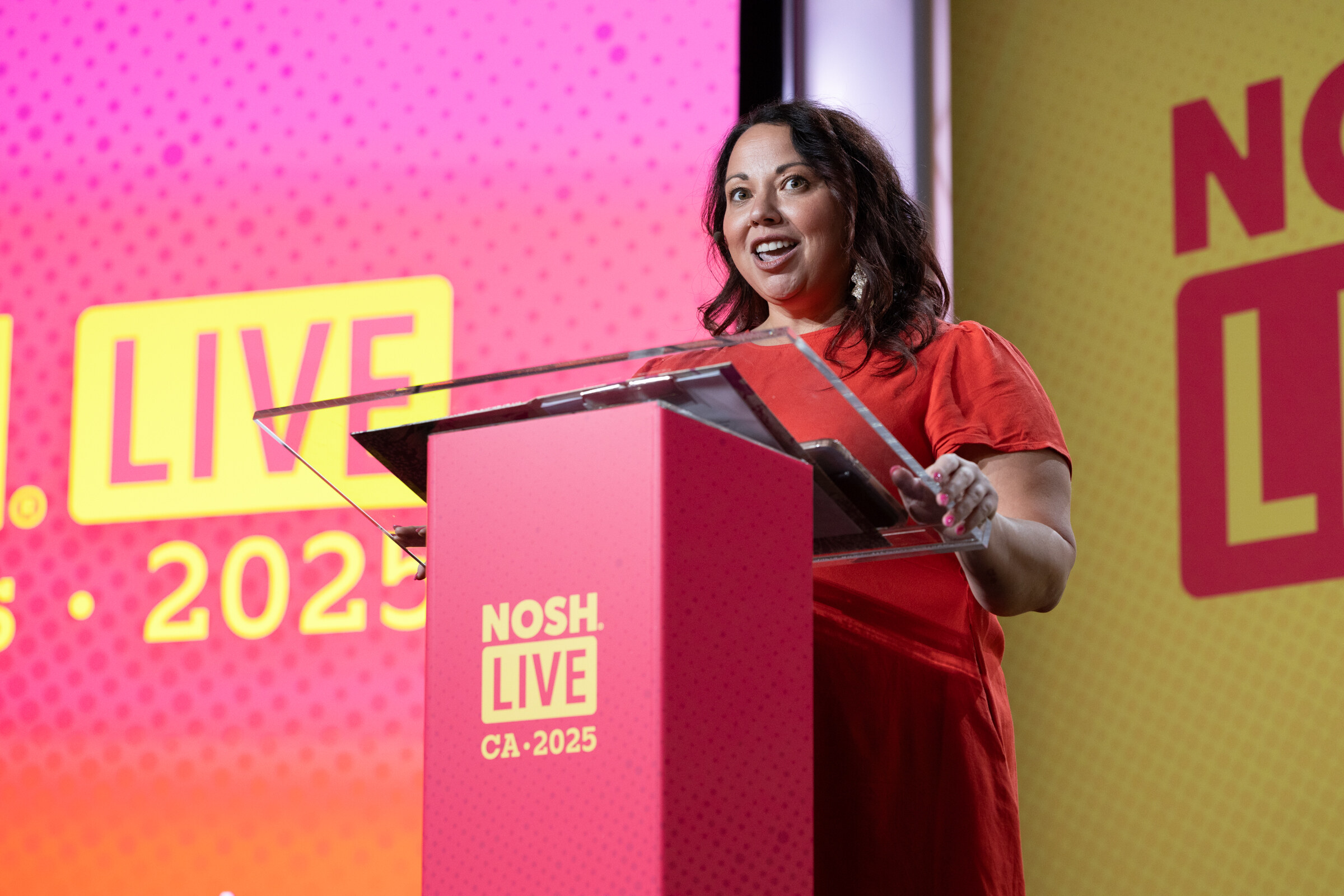 A smiling woman in a red dress at a podium marked Nosh Live CA 2025 against a pink and yellow backdrop.