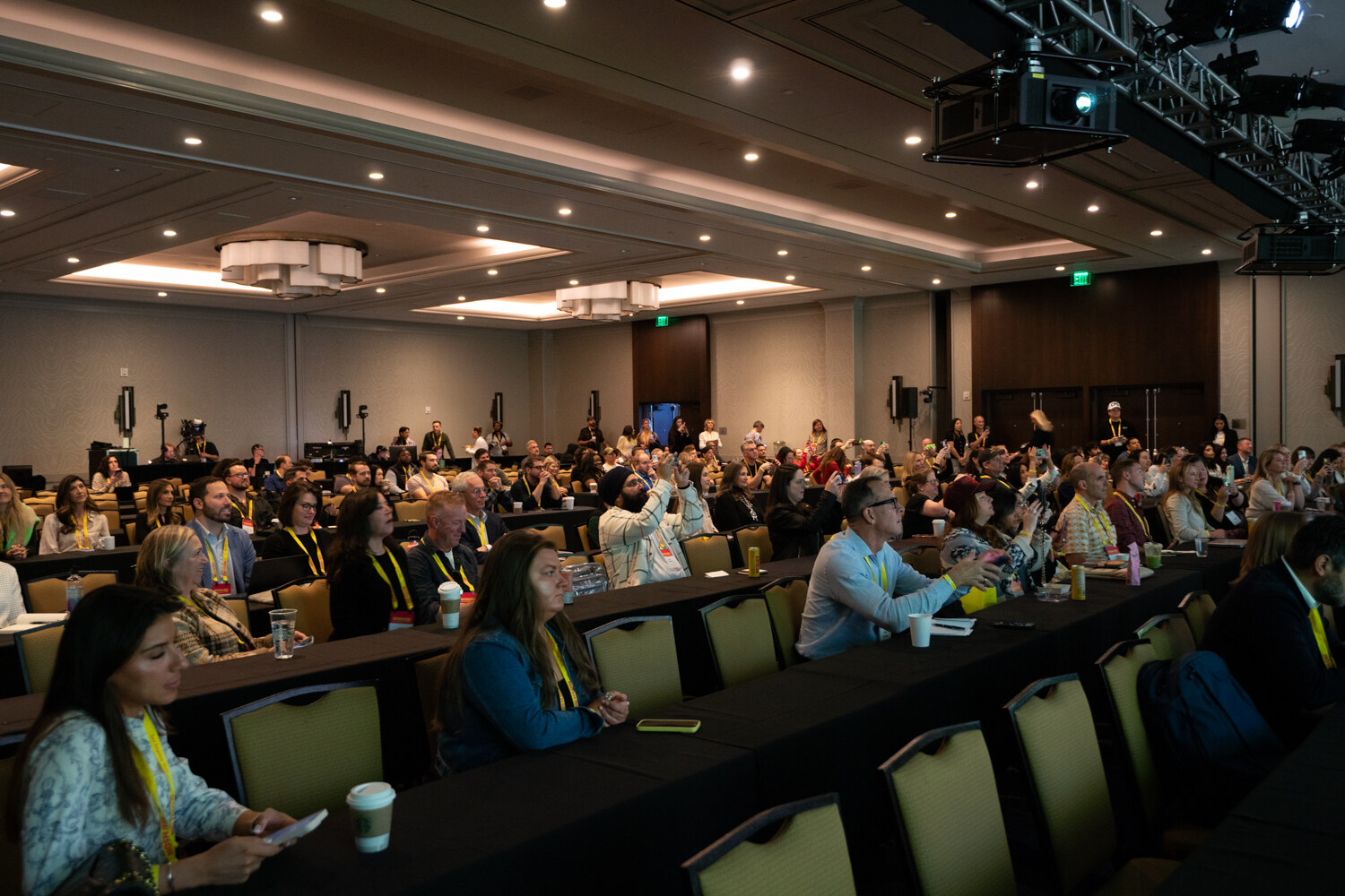 Rows of attendees wearing yellow lanyards seated at long draped tables in a hotel ballroom.