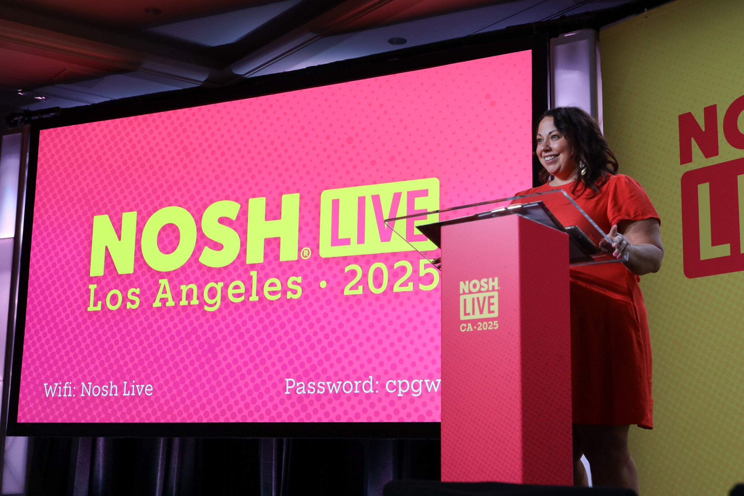 A woman in a red dress speaking at a clear podium beside a pink screen reading Nosh Live Los Angeles 2025 with Wi-Fi details.