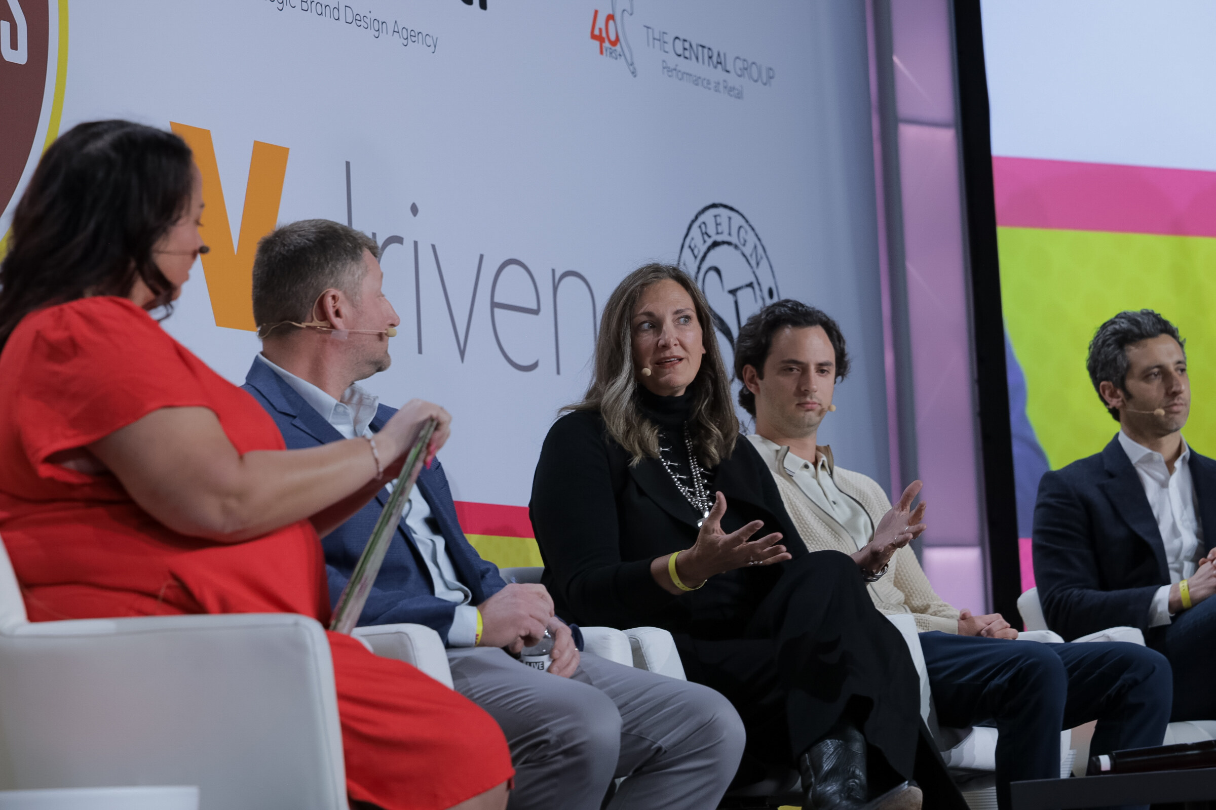 Five people seated in white chairs on stage in front of a Vdriven banner, with the moderator in a red dress at left.