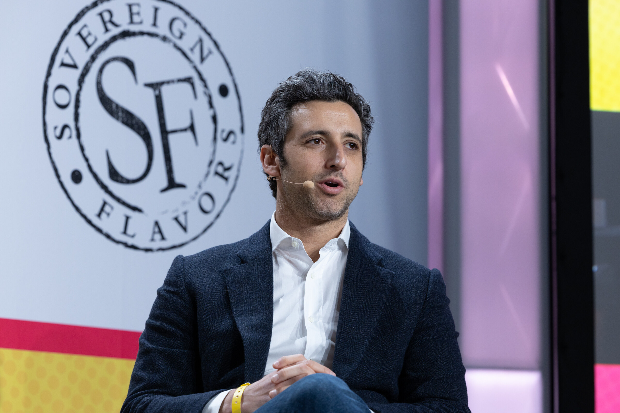 A man in a navy blazer with a headset microphone speaking in front of a Sovereign Flavors logo backdrop.