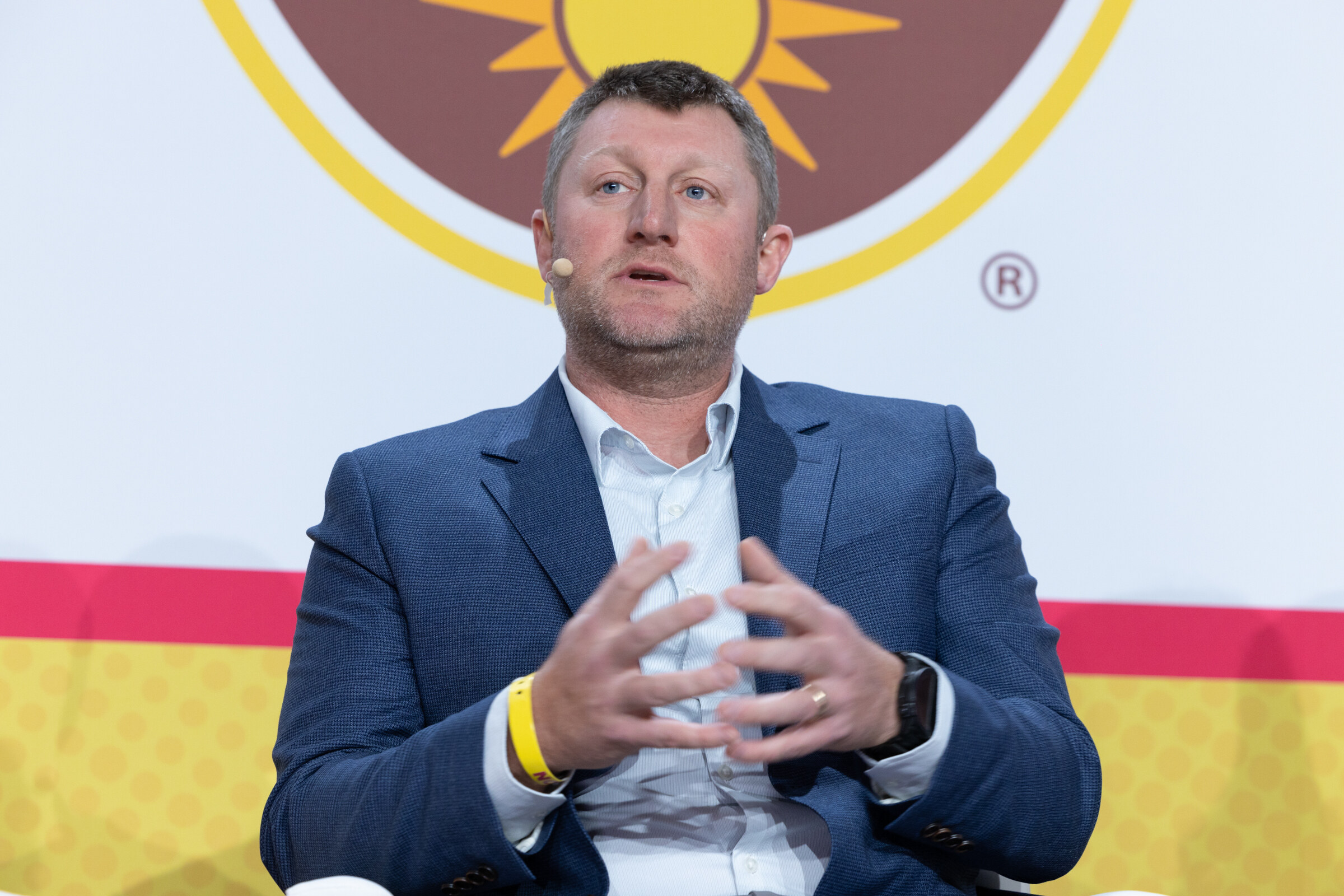A man in a blue blazer with a headset microphone holding his hands together in front of a California Walnuts backdrop.