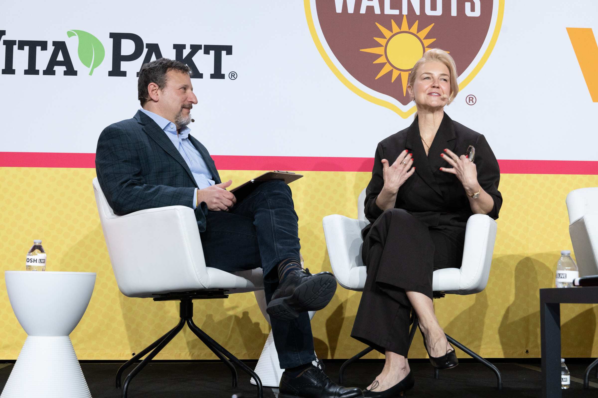 A man with a clipboard and a woman in a black outfit seated in white chairs on stage under a Vita-Pakt sign.