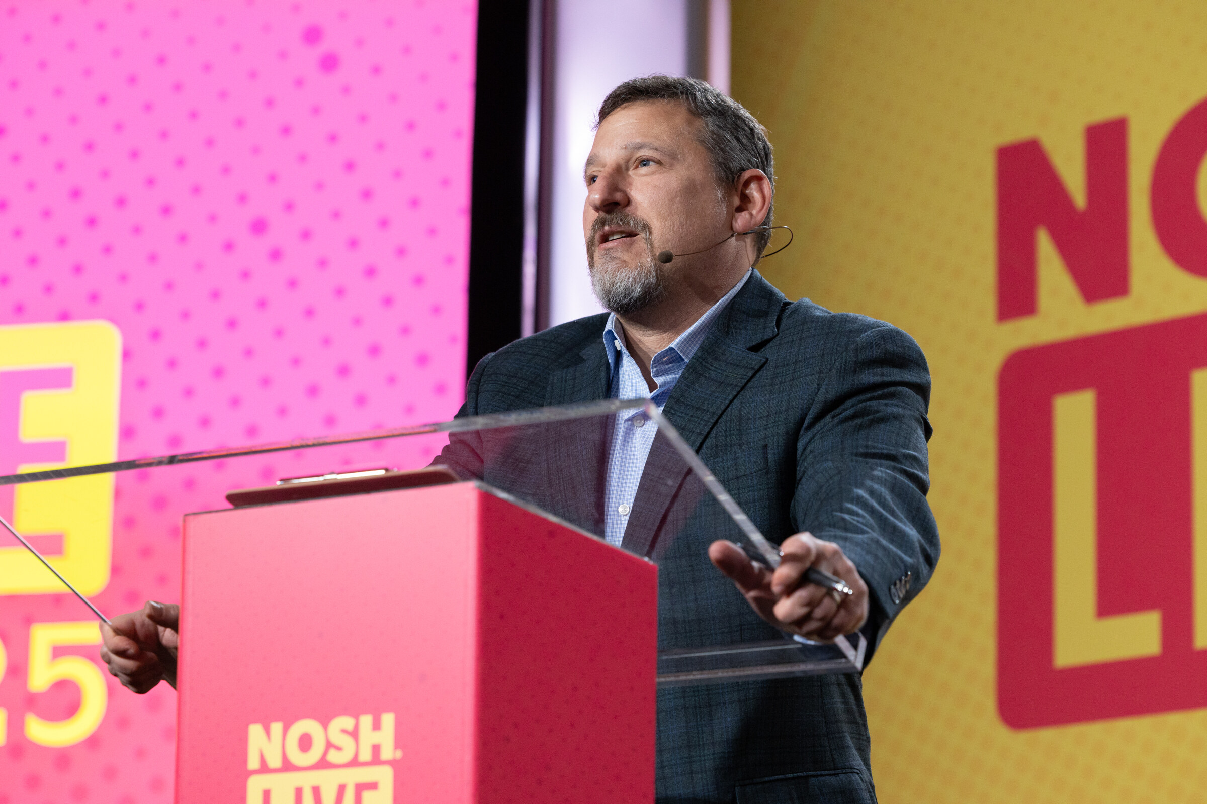 A man in a dark blazer with a headset microphone speaking at a podium in front of a pink and yellow Nosh Live backdrop.