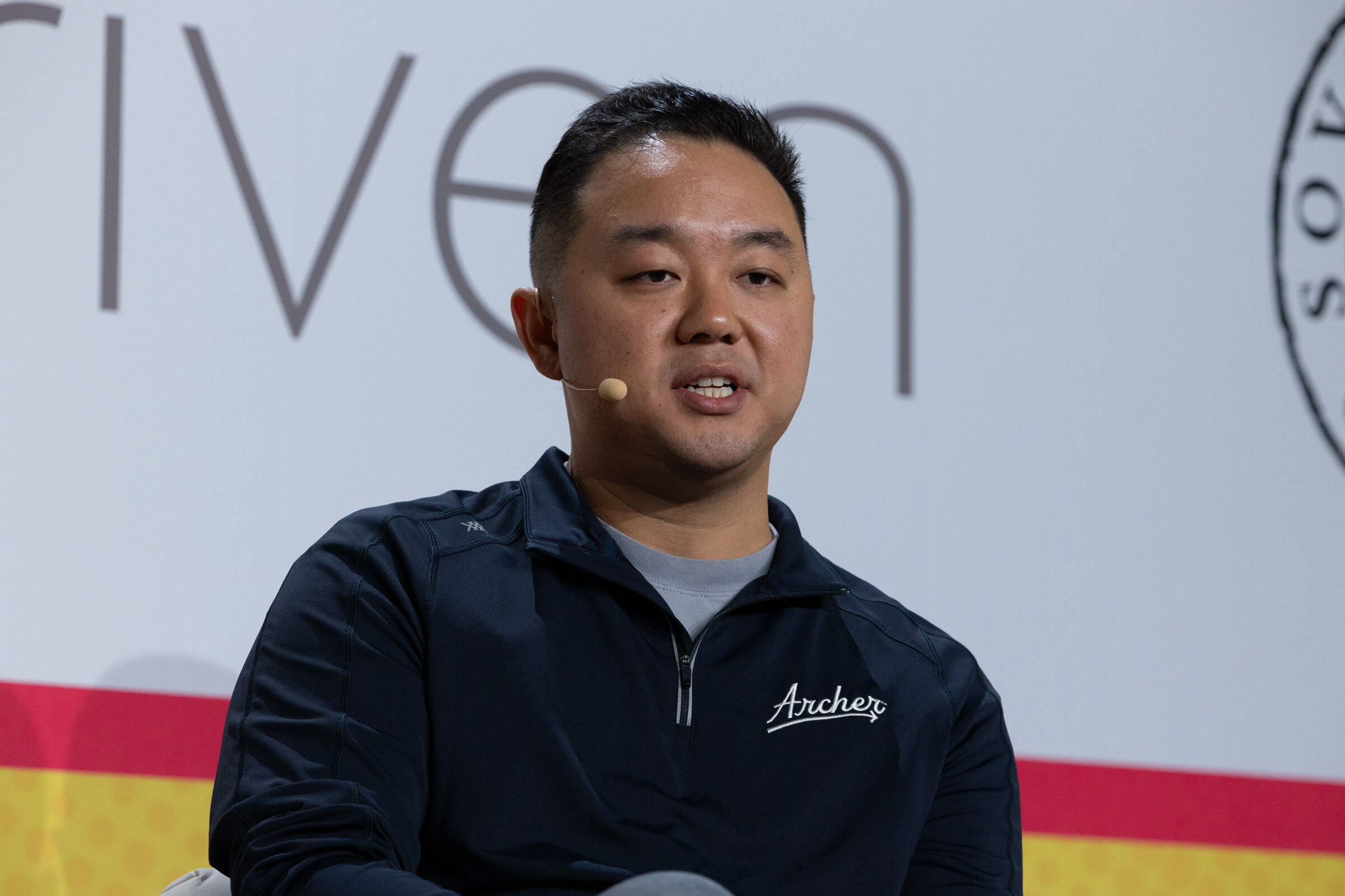A man in a navy pullover with an Archer logo speaking into a headset microphone.