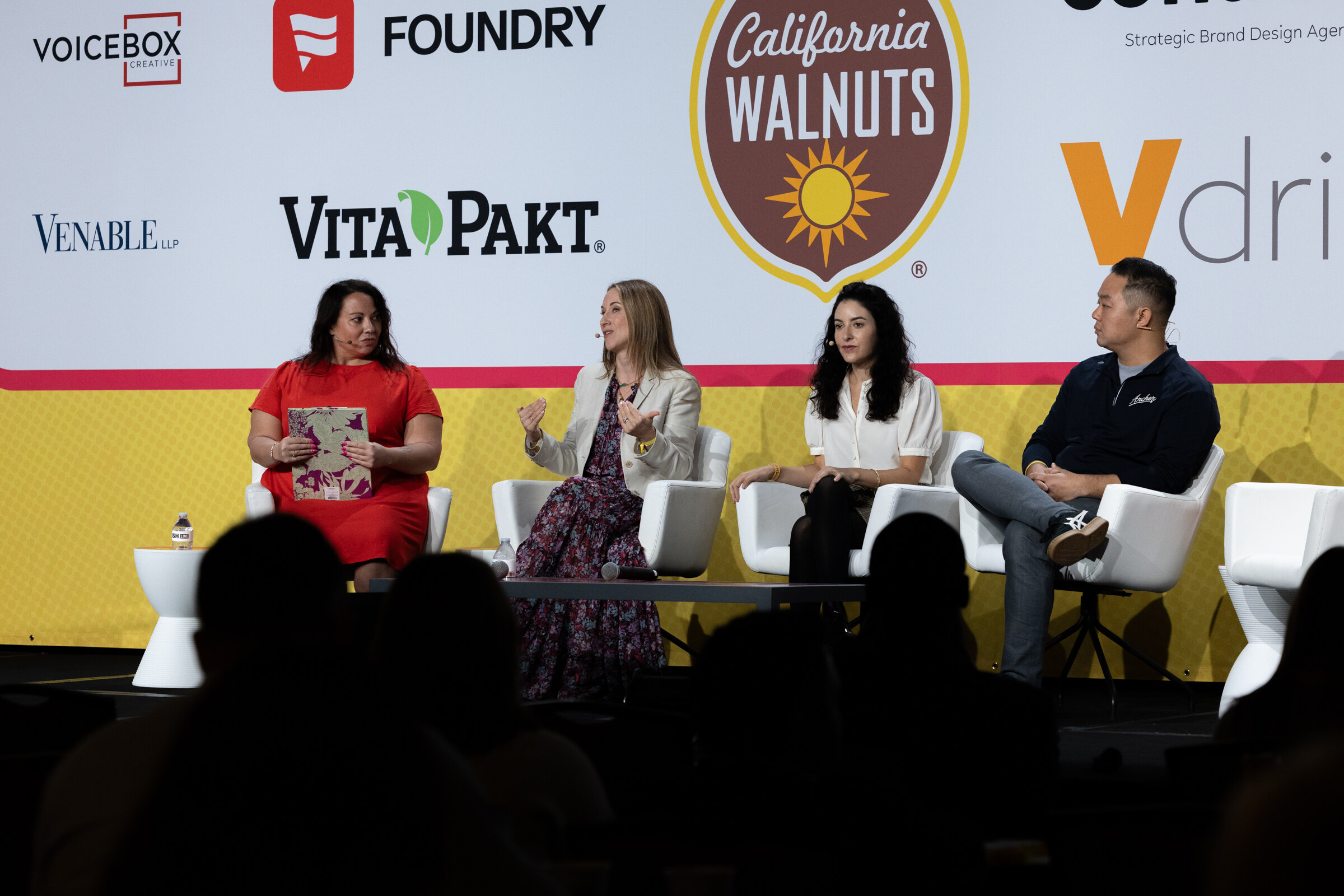 Four people seated on a stage in front of a banner with sponsor logos, the moderator in a red dress at left.