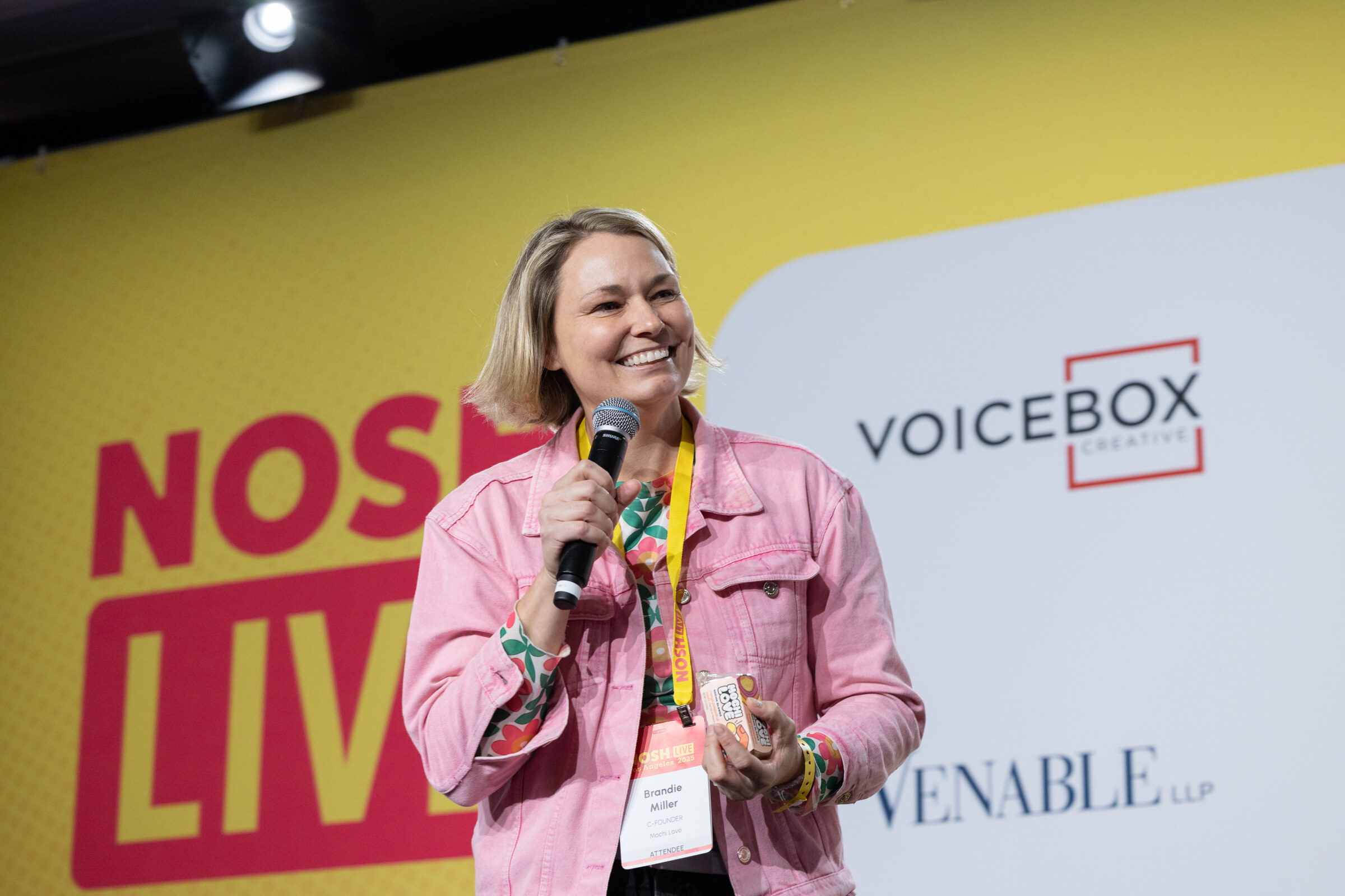 A woman in a pink denim jacket holding a microphone and a small product in front of a Nosh Live and Voicebox backdrop.