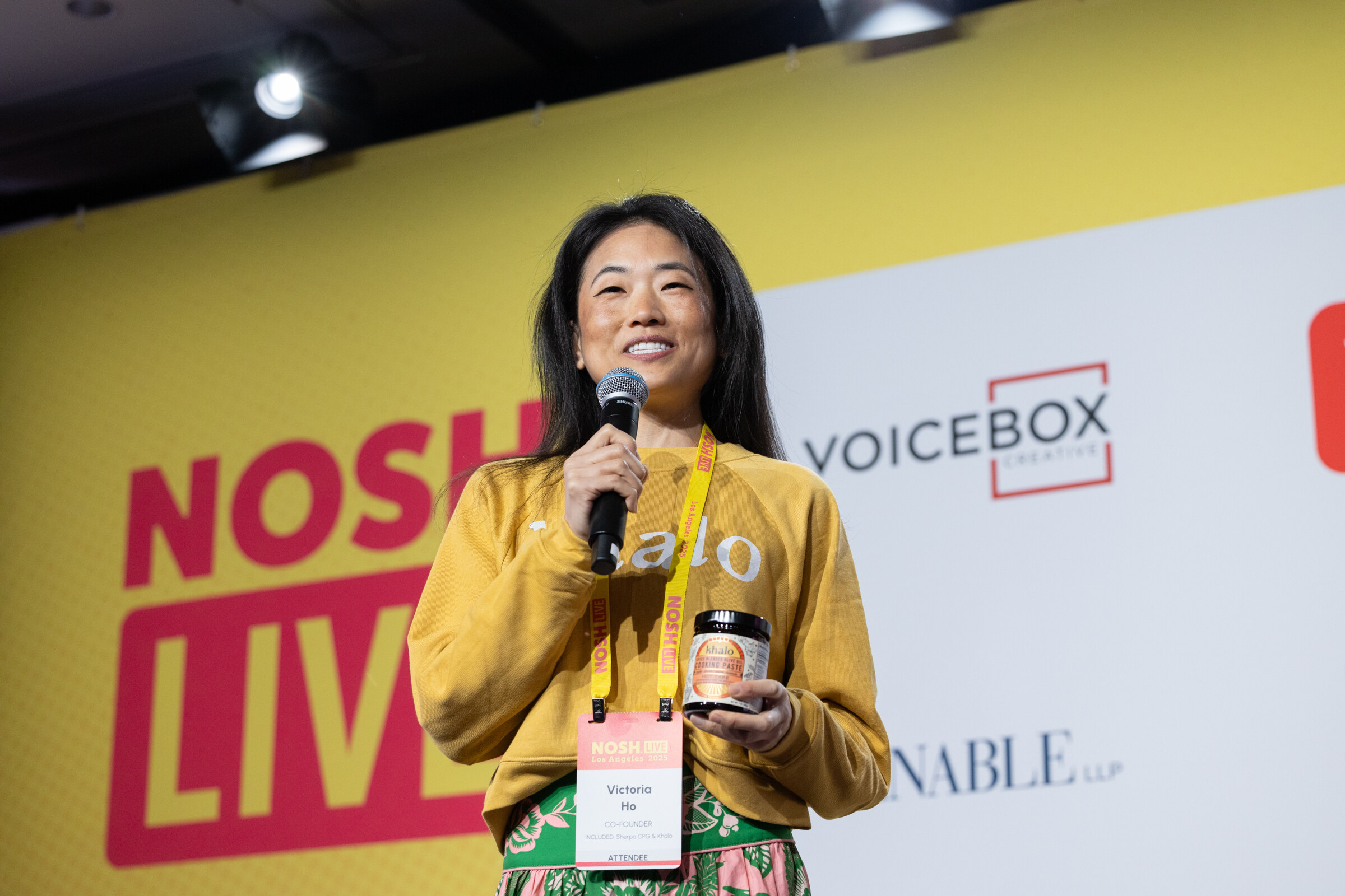A woman in a mustard sweatshirt speaking into a microphone while holding a jar of product.