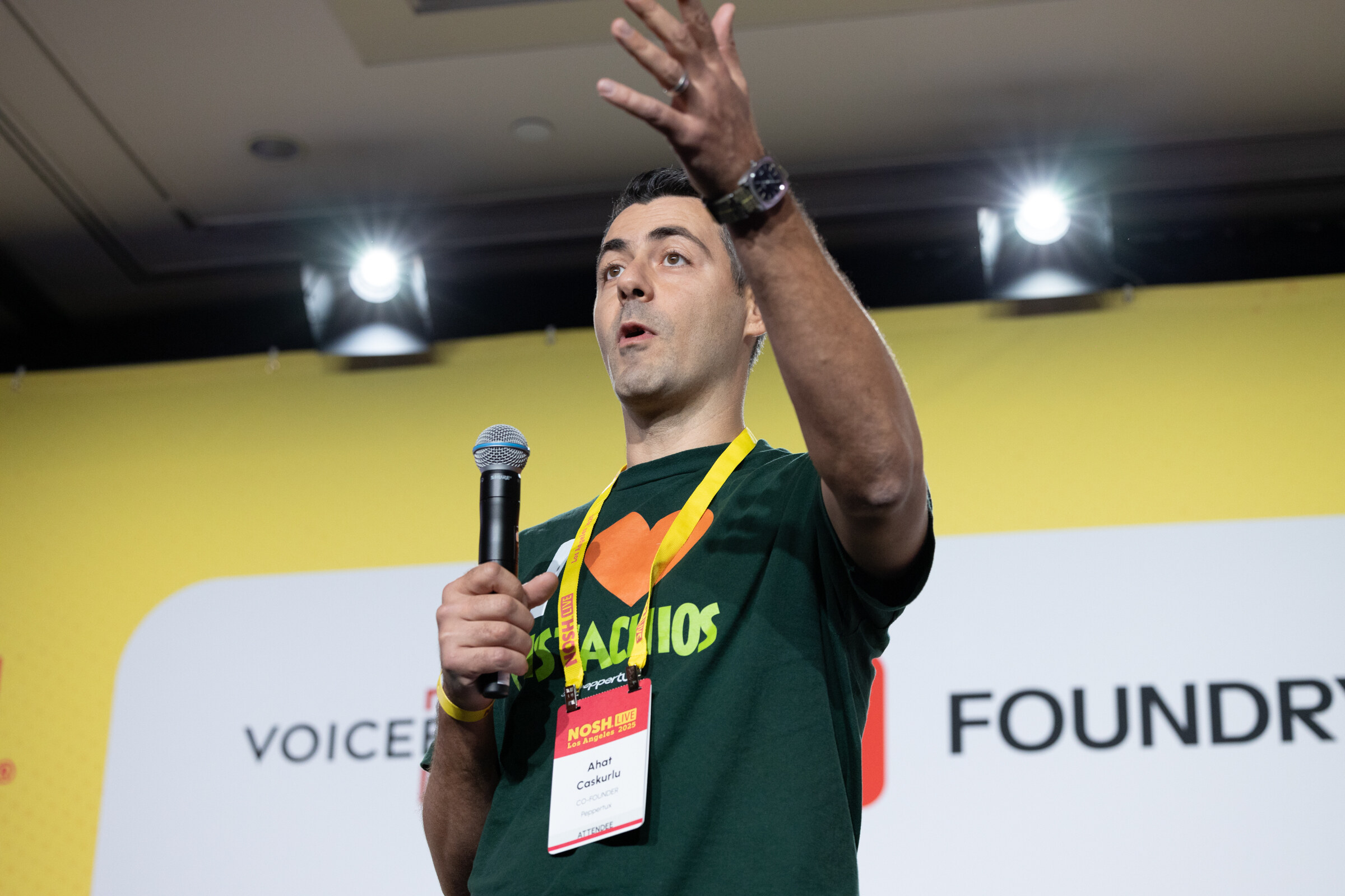 A man in a dark green shirt holding a microphone and raising one arm while speaking on stage.
