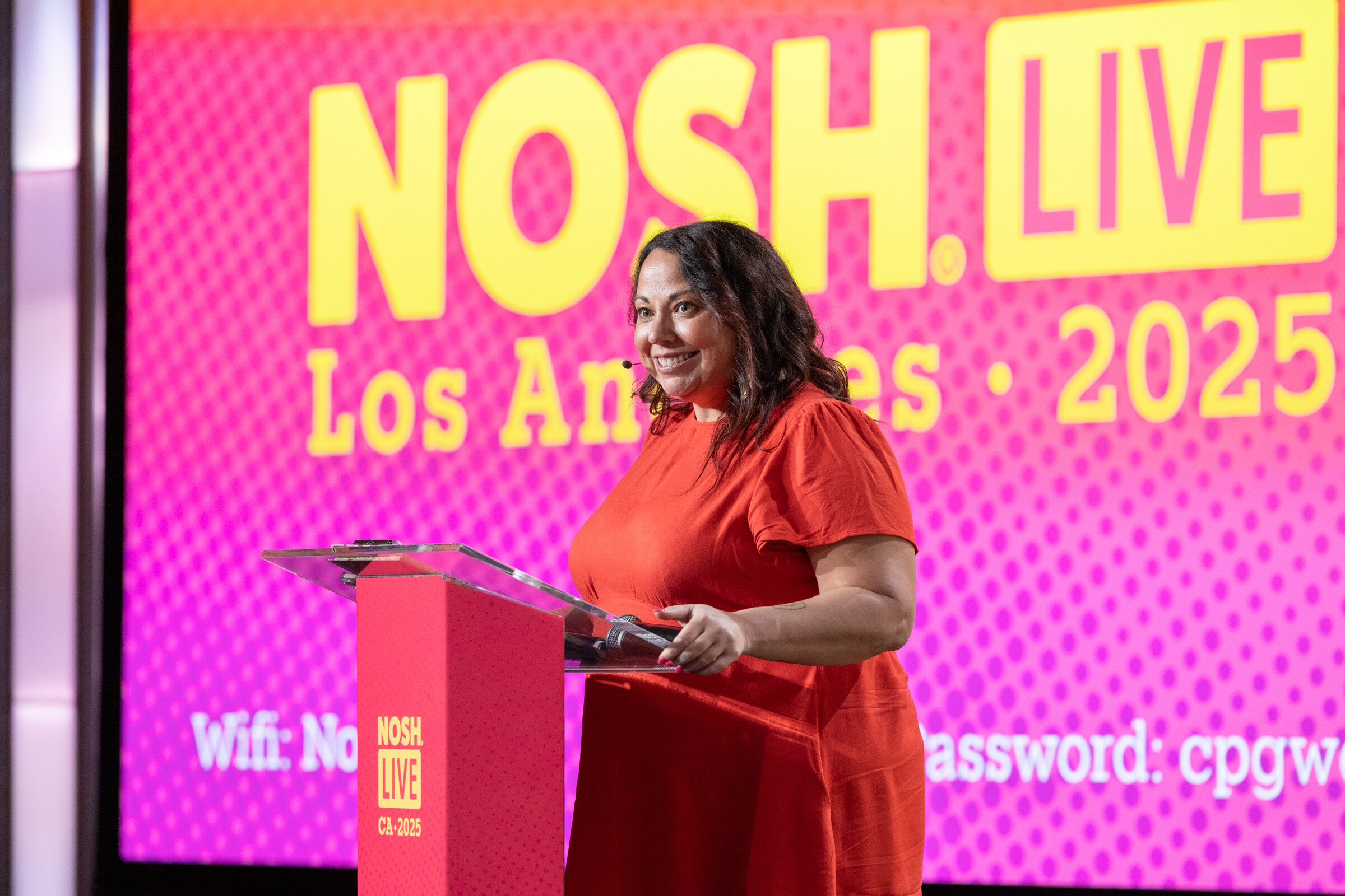 A woman in an orange-red dress at a podium in front of a pink Nosh Live Los Angeles 2025 screen.