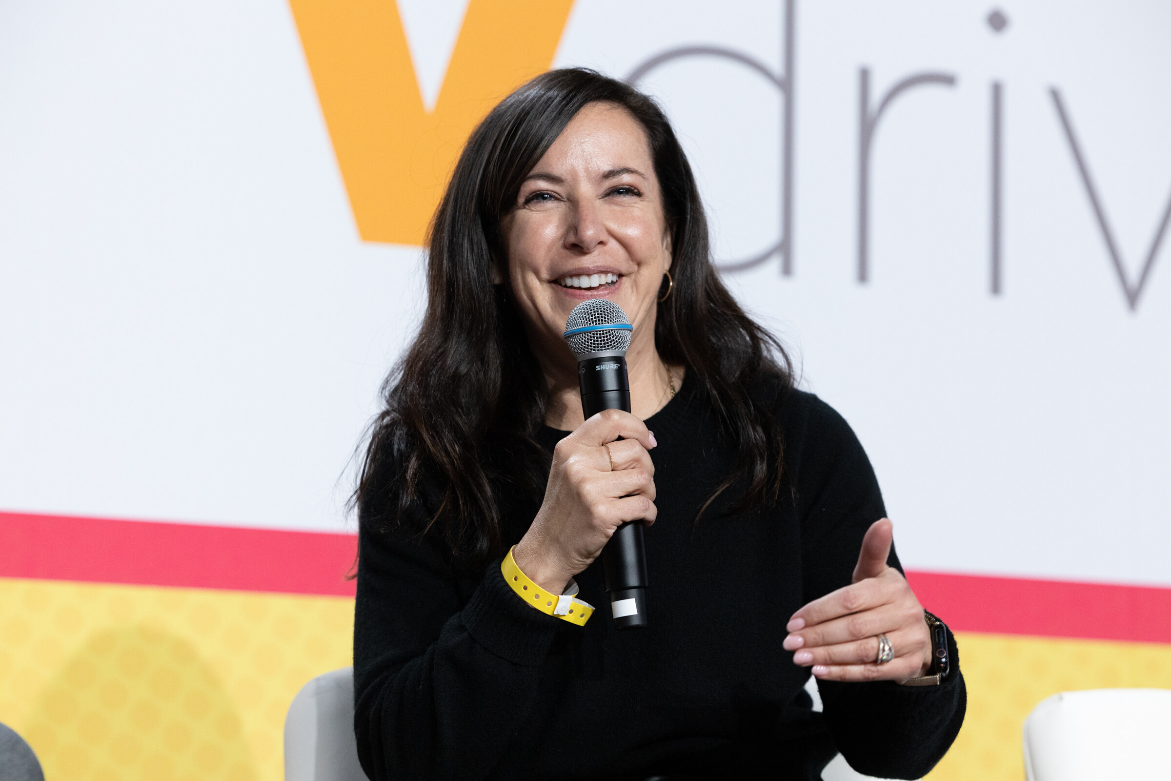 A woman in a black sweater holding a microphone and gesturing, seated in front of a Vdriven backdrop.
