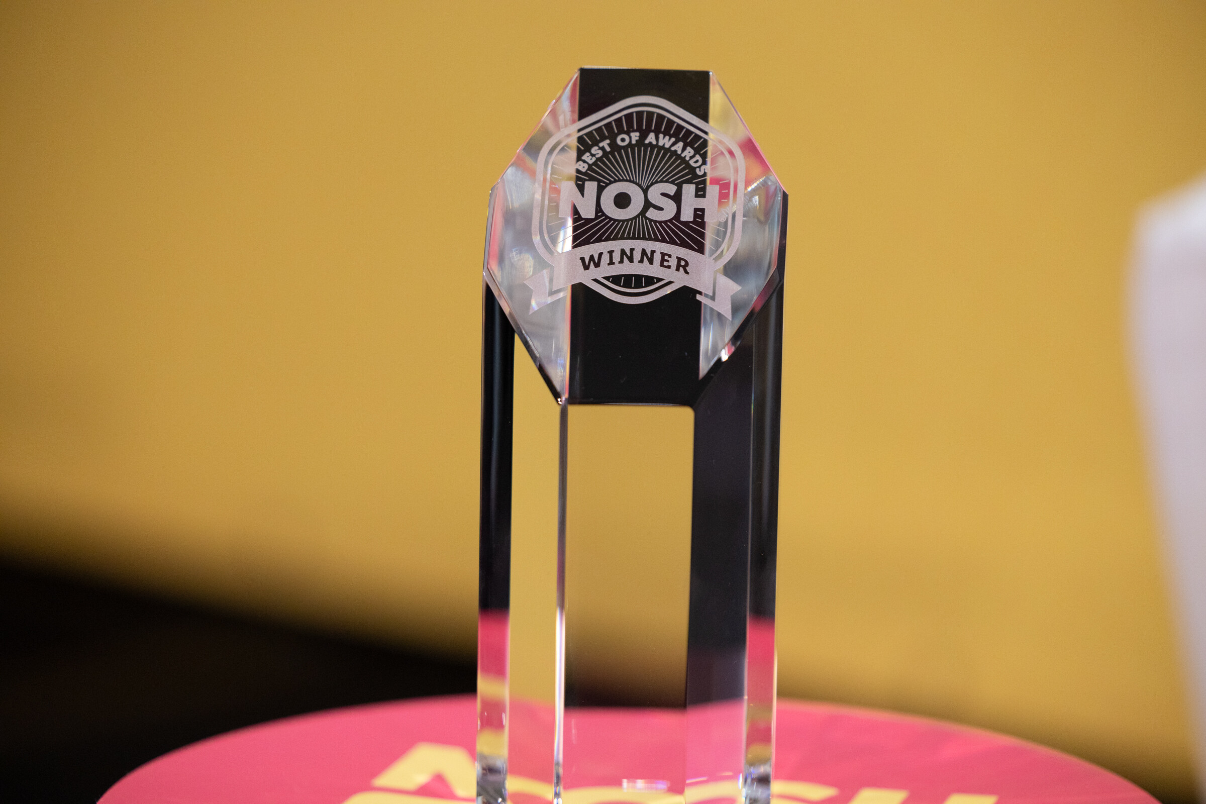 A clear glass trophy engraved Nosh Best of Awards Winner on a yellow stage backdrop.