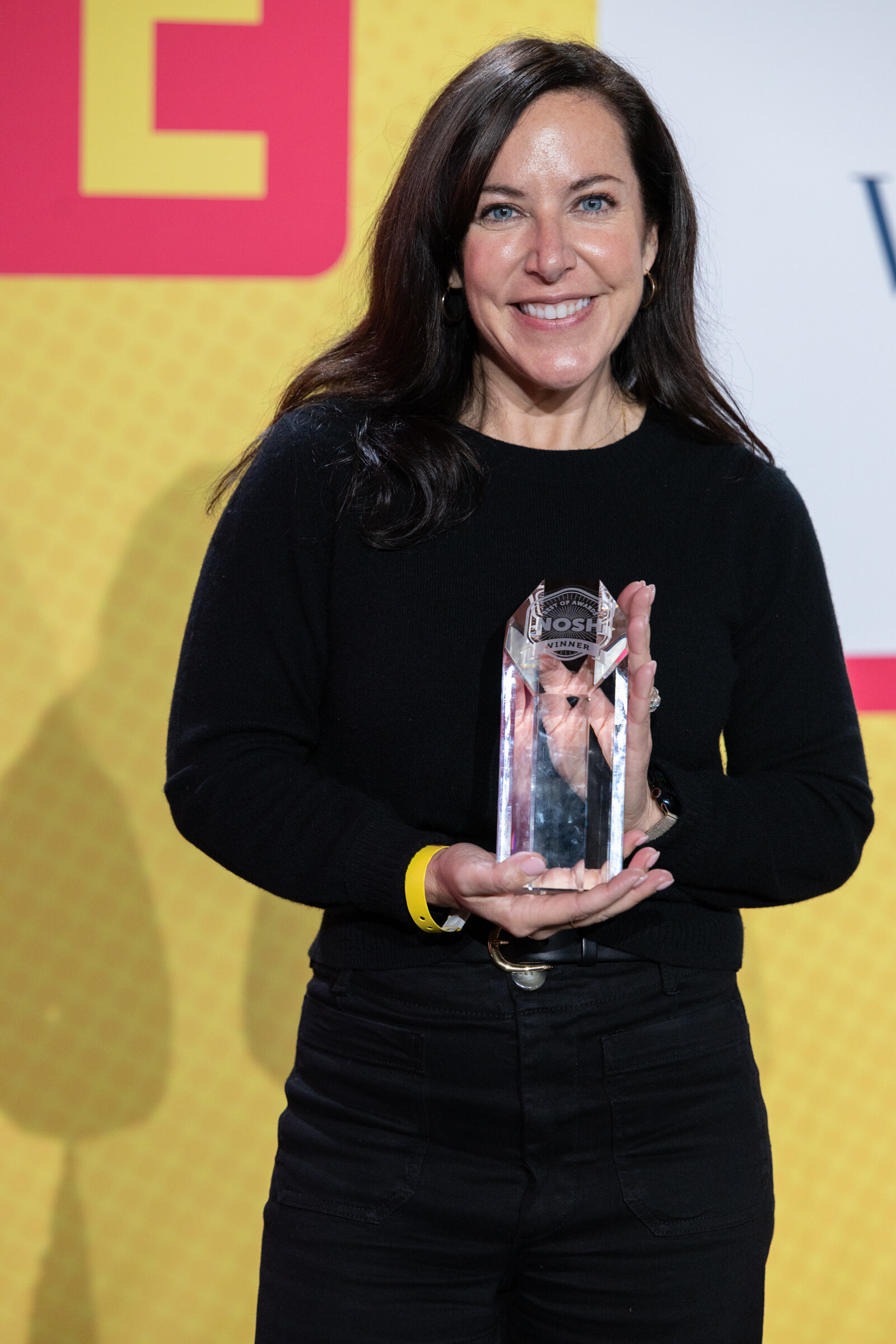 A smiling woman in black holding a glass trophy against a yellow backdrop.