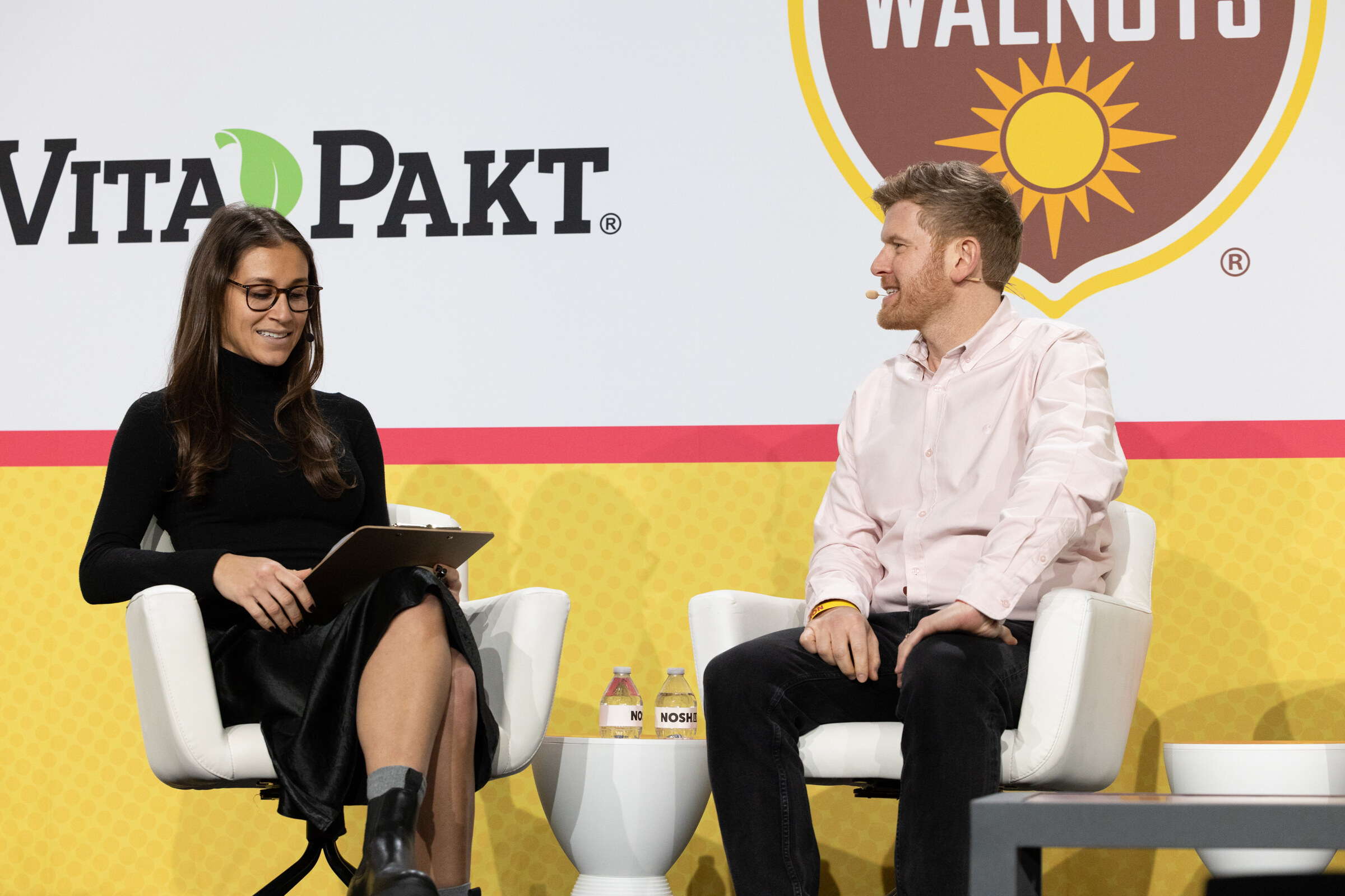 A woman in black with a tablet and a man in a light pink shirt seated on white chairs under Vita-Pakt and California Walnuts signs.