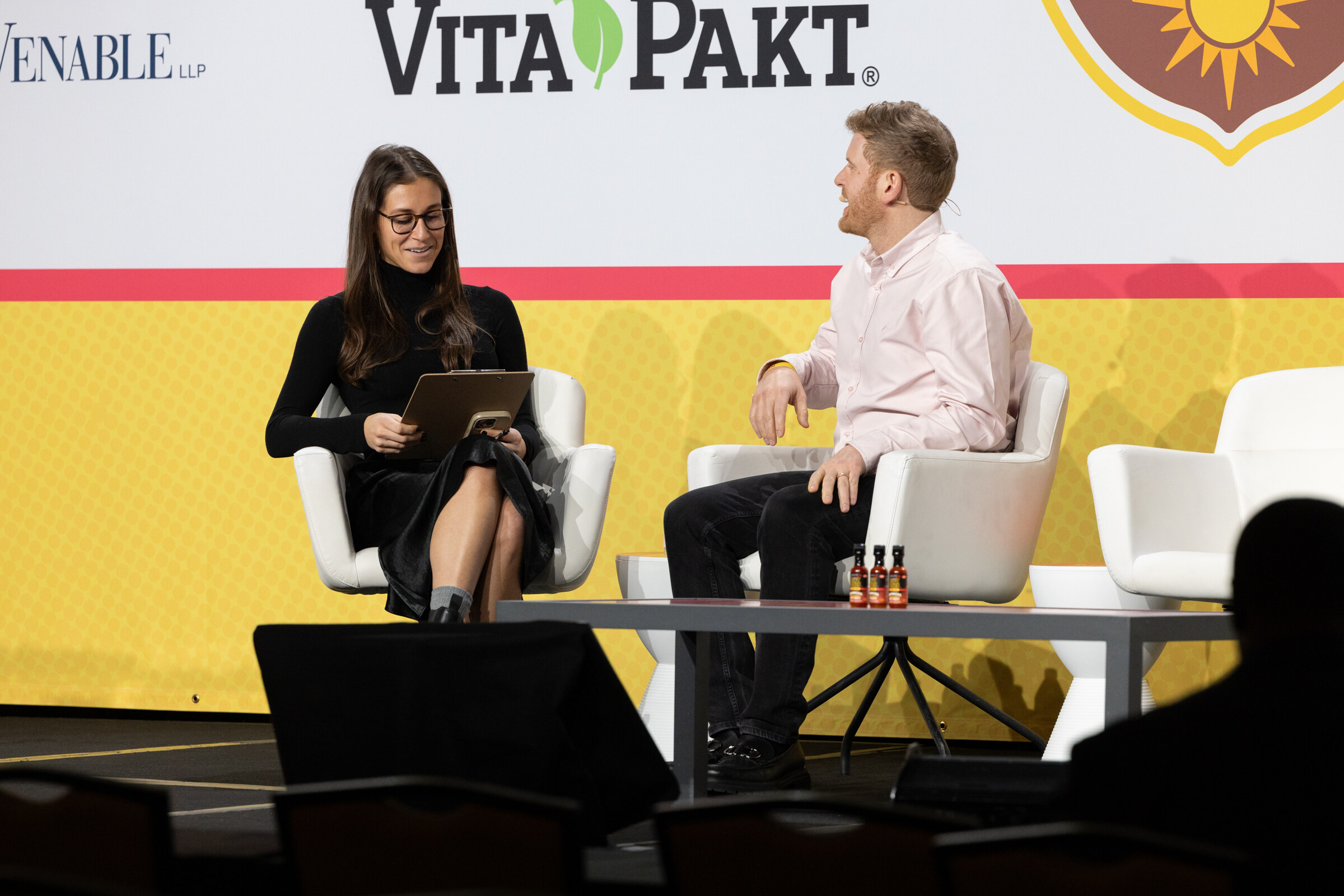 Two people seated on stage, a woman holding a tablet and a man in a pink shirt, with small bottles on a low table.