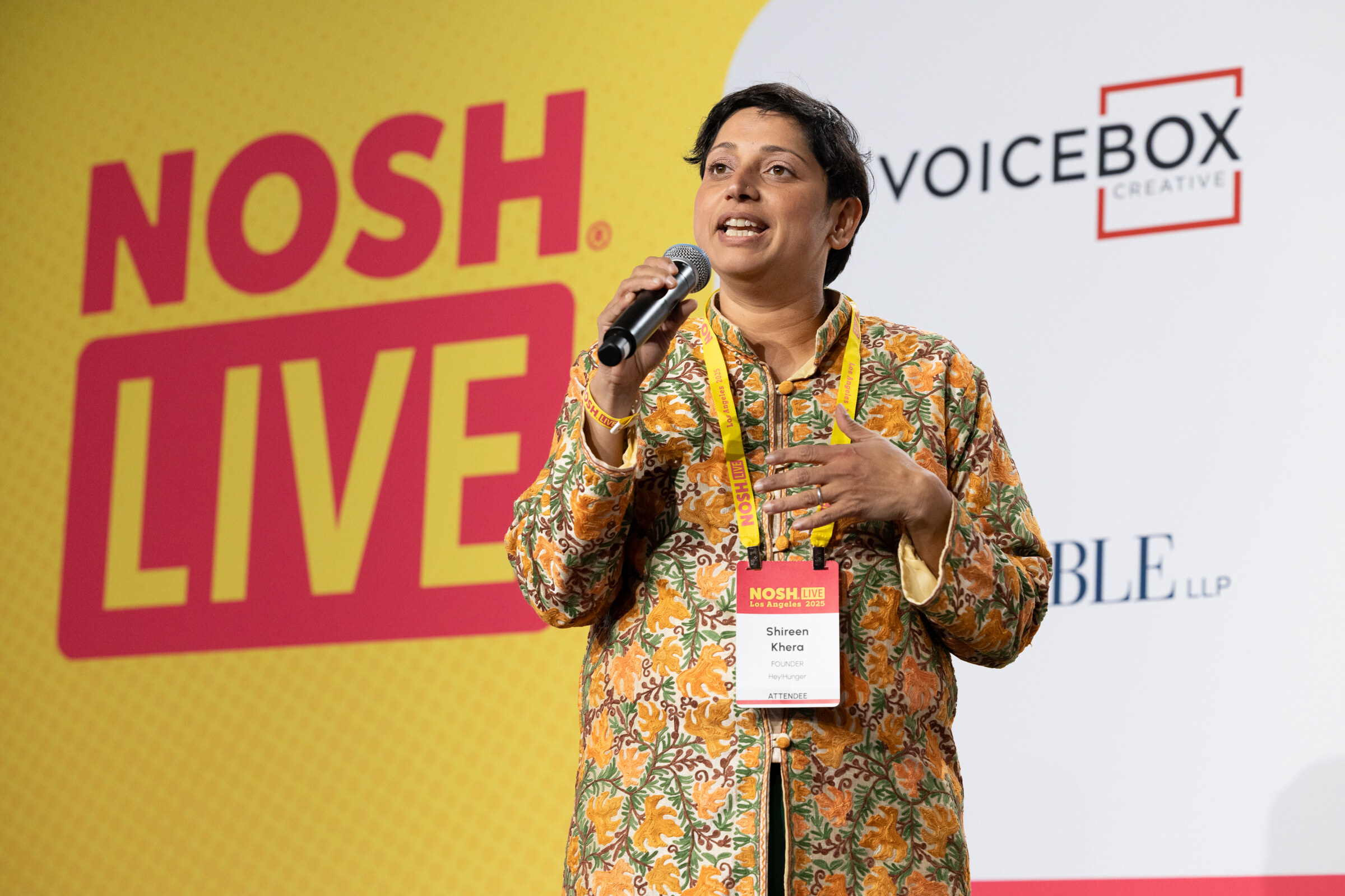 A woman in a printed orange jacket speaking into a handheld microphone on stage.