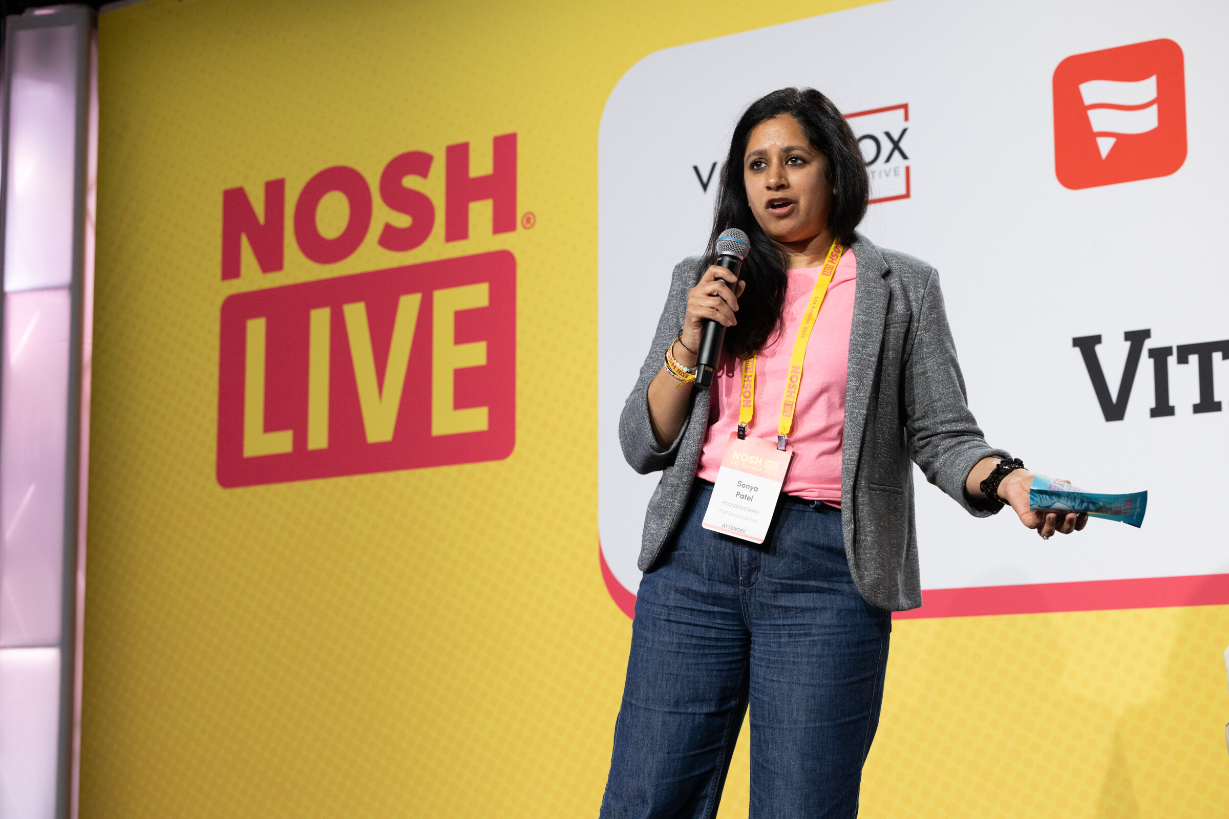 A woman in a gray blazer and pink top speaking into a microphone while holding a blue snack bar.