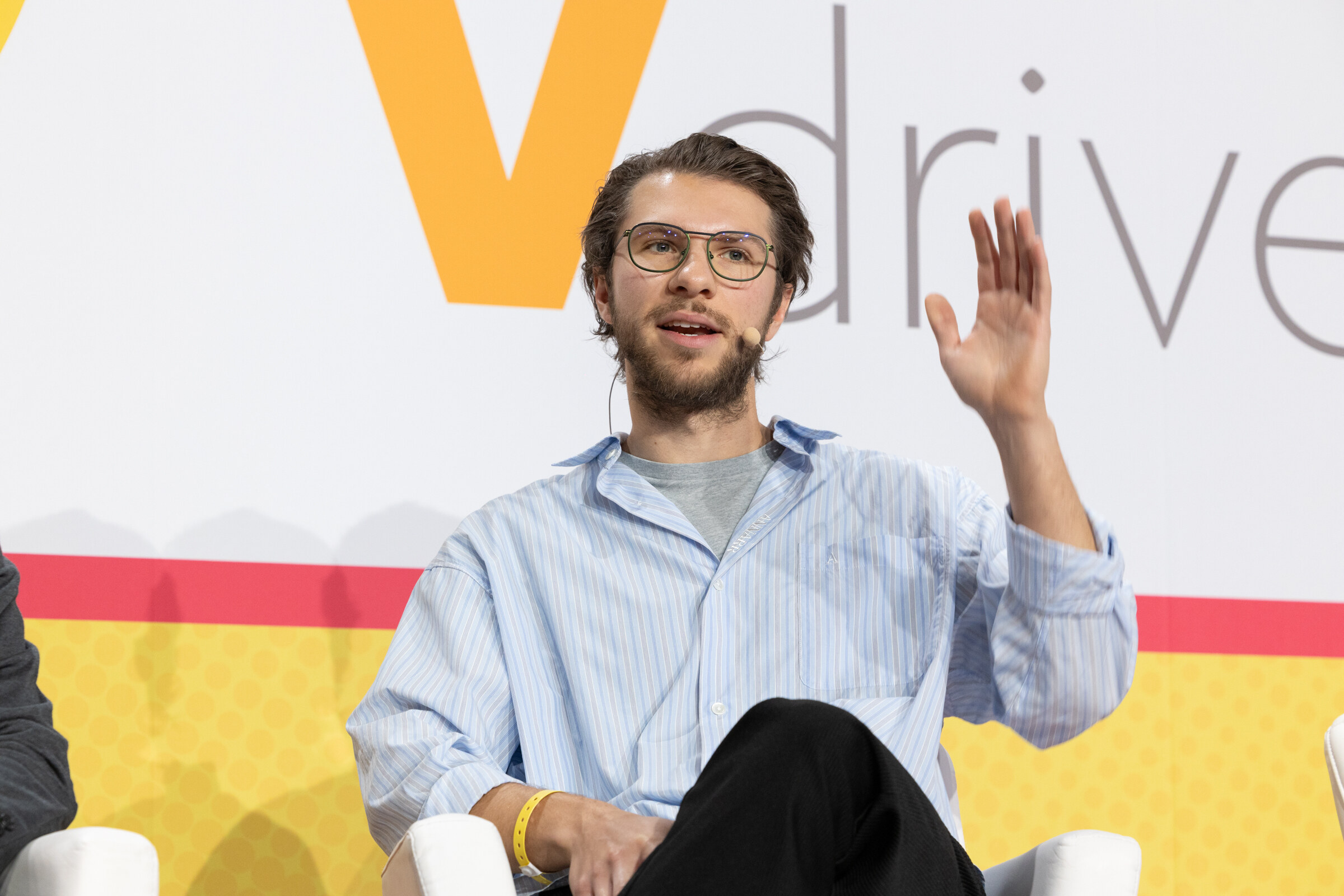 A bearded man in a light blue striped shirt raising one hand while speaking, in front of a Vdriven backdrop.