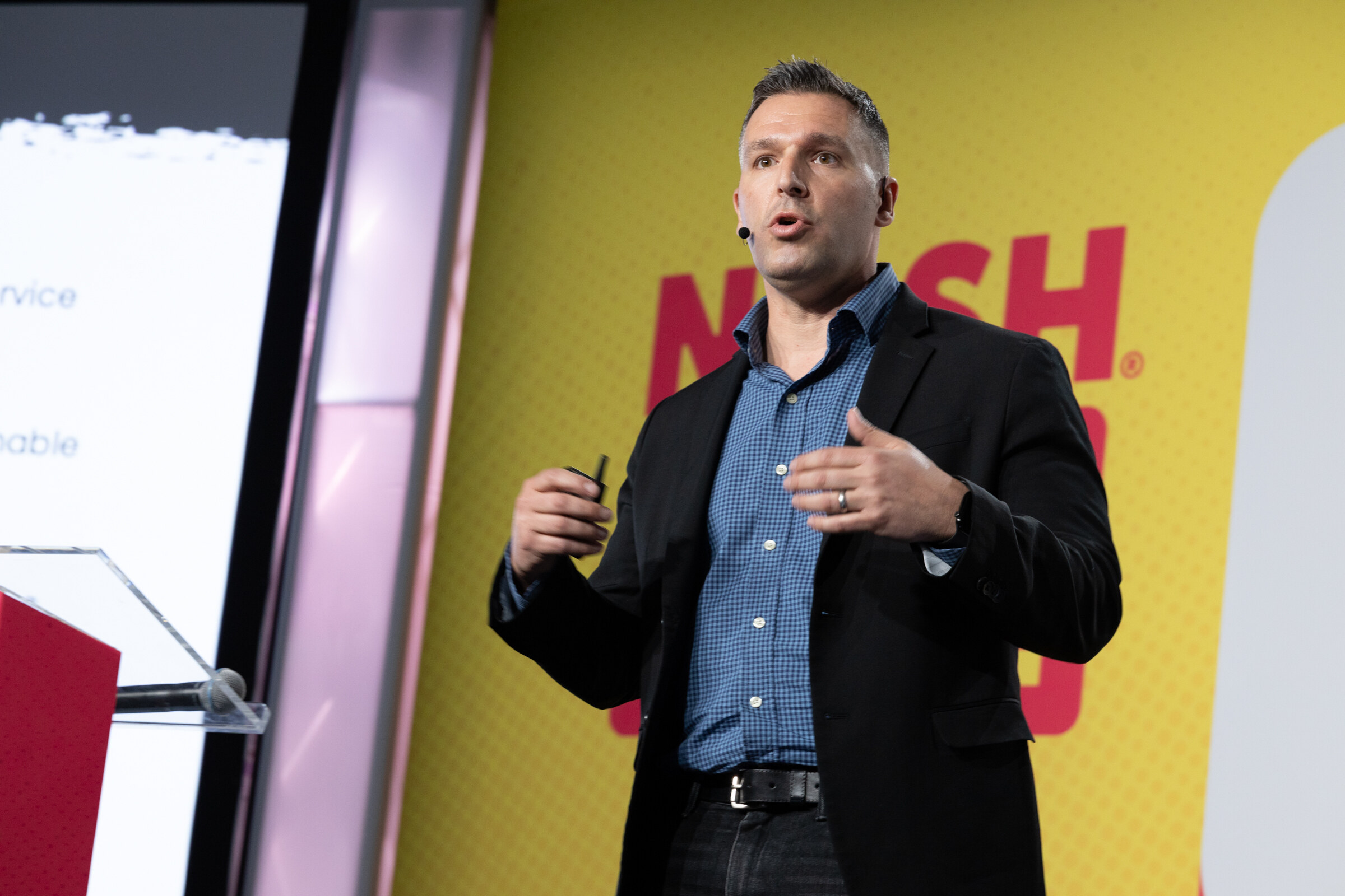 A man in a dark blazer gesturing with both hands on stage next to a clear podium.