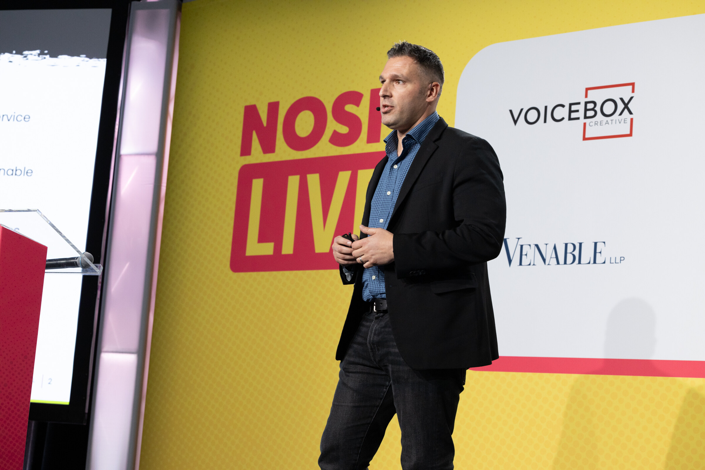A man in a dark blazer and blue checked shirt speaking on stage in front of a Nosh Live and Voicebox backdrop.
