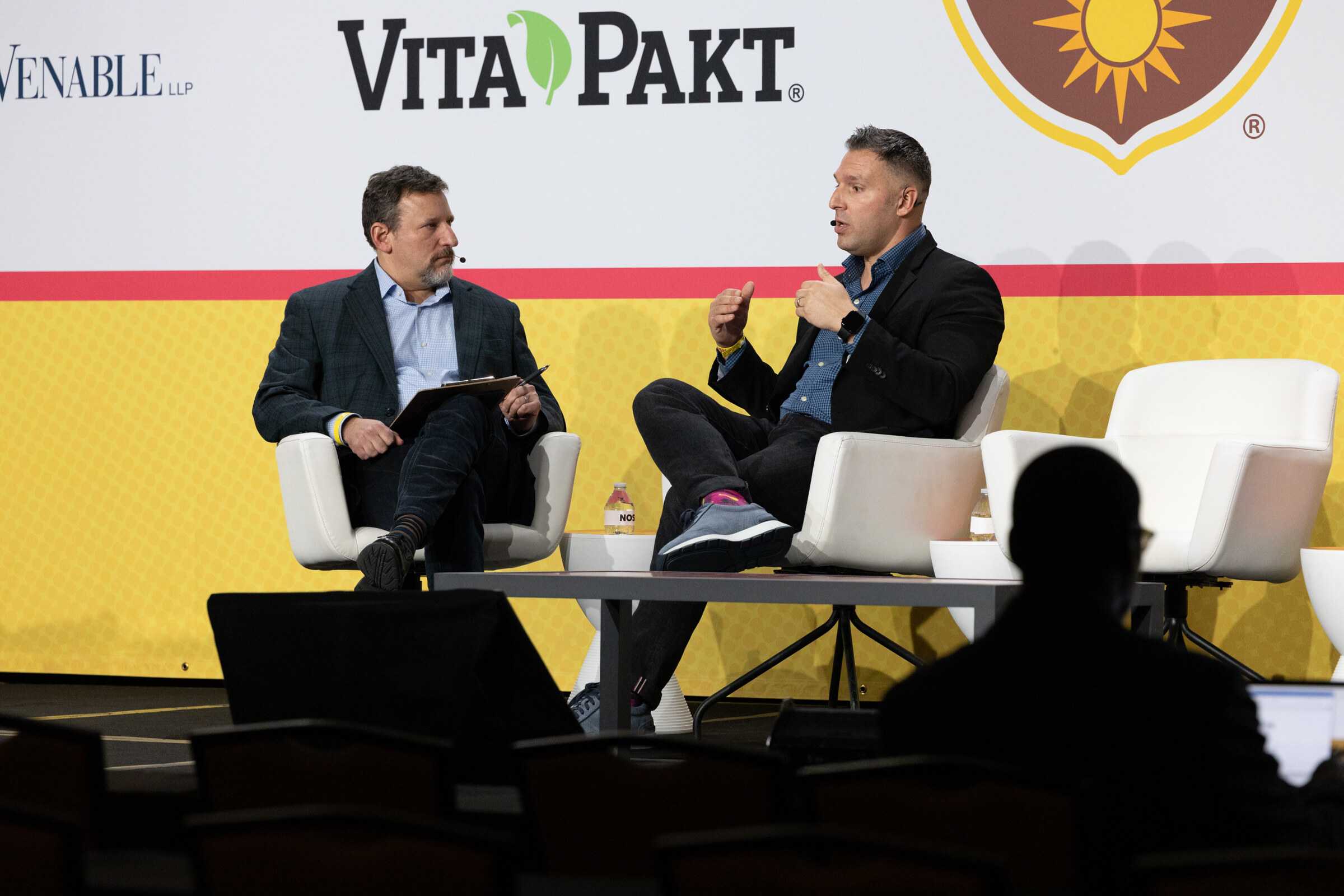 Two men seated in white chairs on stage in front of a Vita-Pakt sign, one speaking and gesturing.