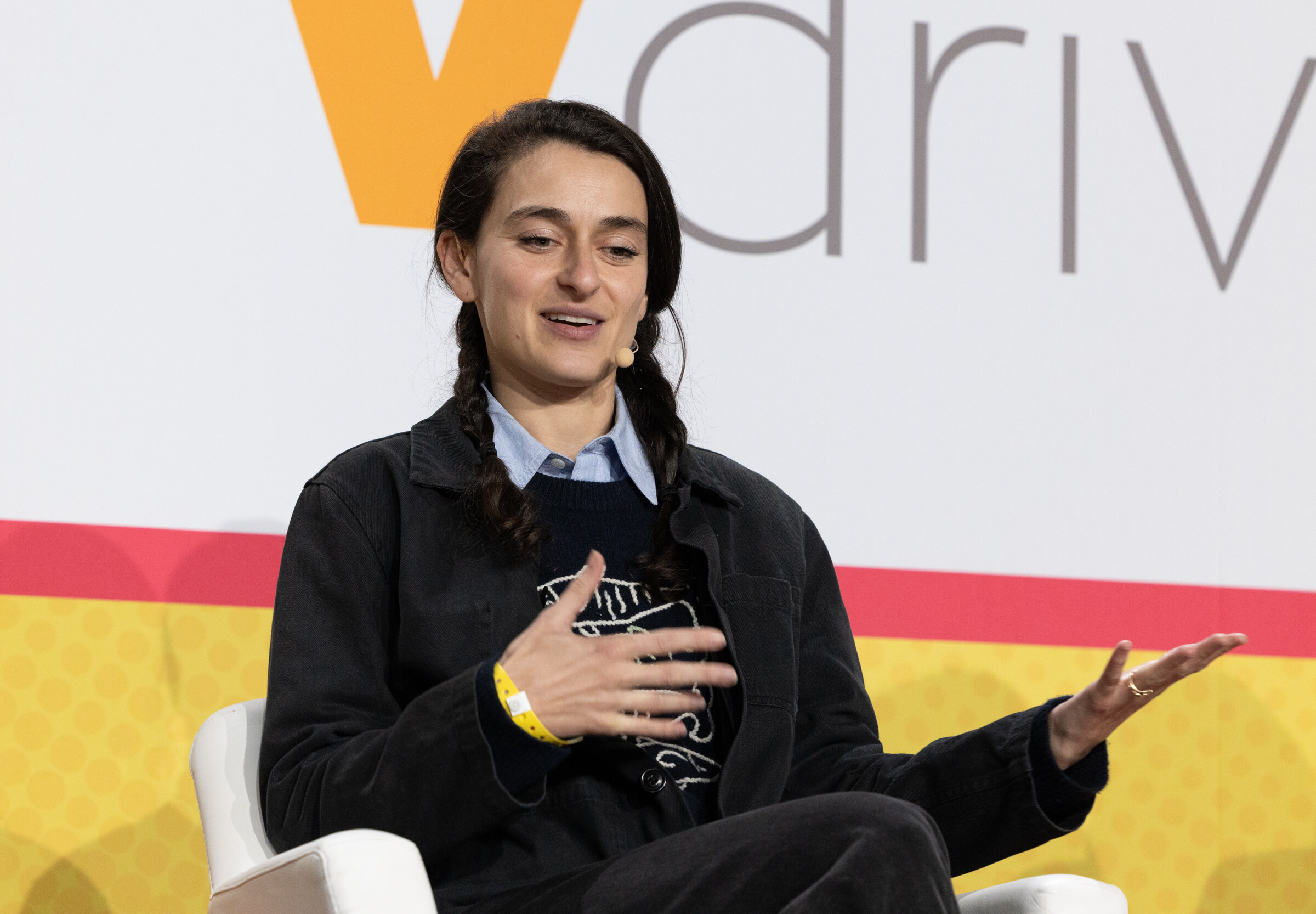 A woman with braids in a dark jacket gesturing with both hands in front of a Vdriven logo.