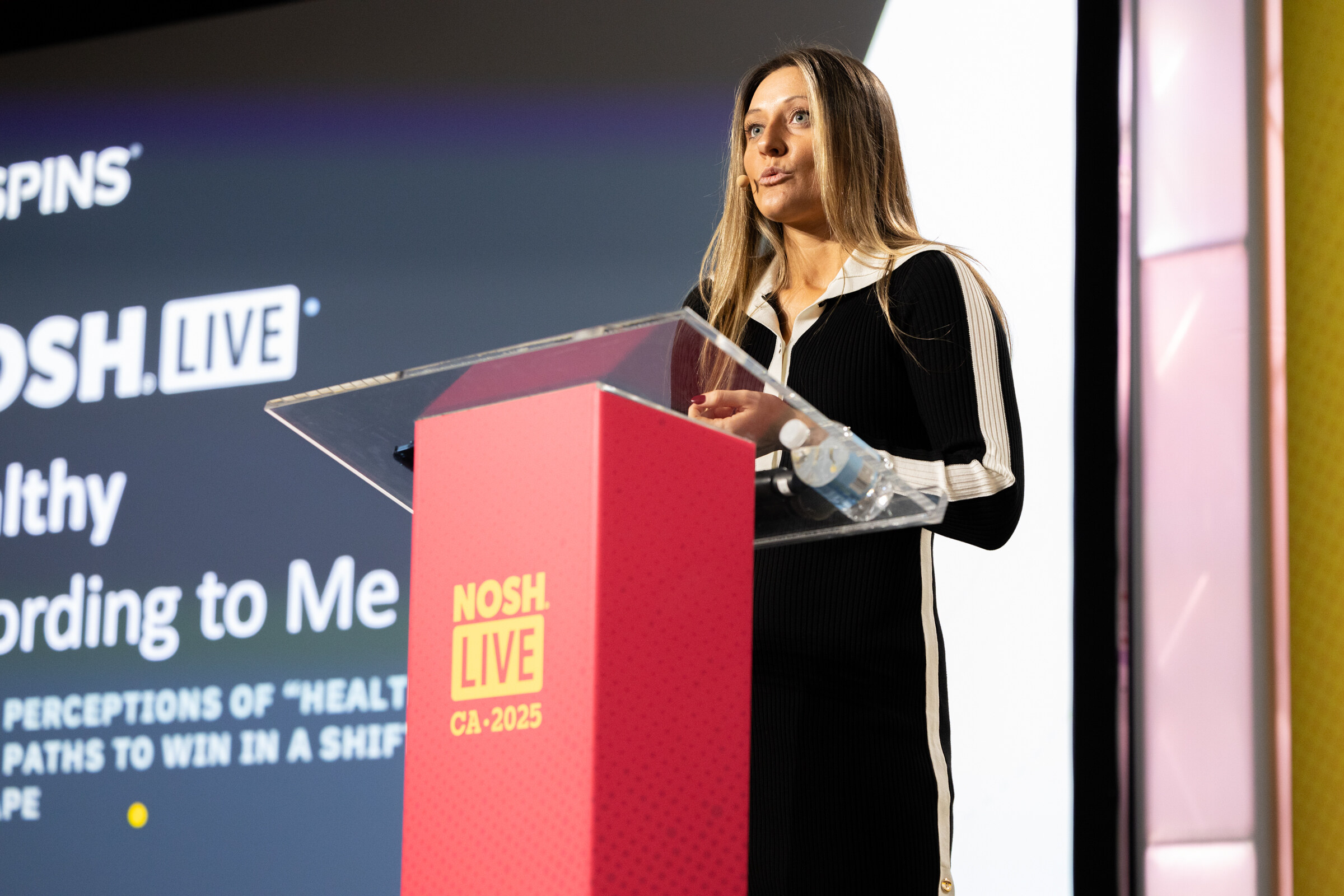 A woman in a black and cream dress at a podium in front of a screen reading Healthy According to Me with a SPINS logo.