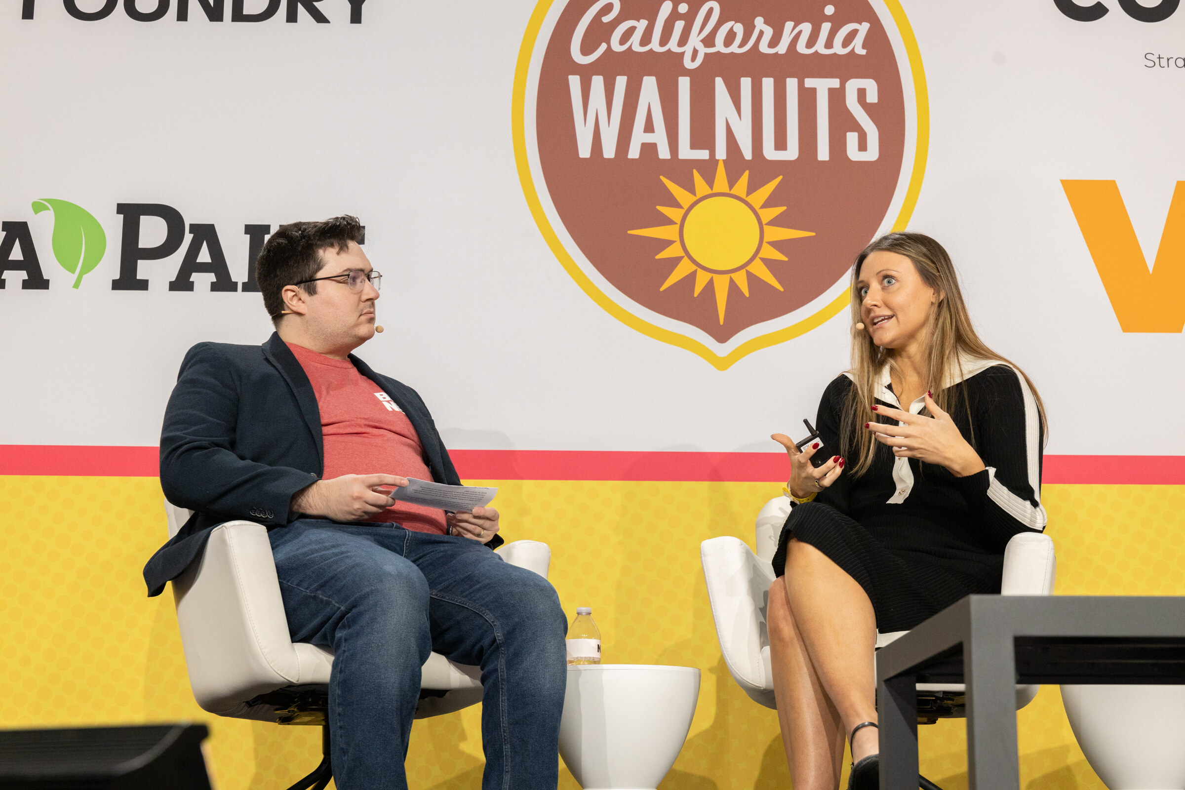 A man in a navy blazer and red shirt seated across from a woman in a black dress on stage in front of California Walnuts and Vdriven signs.