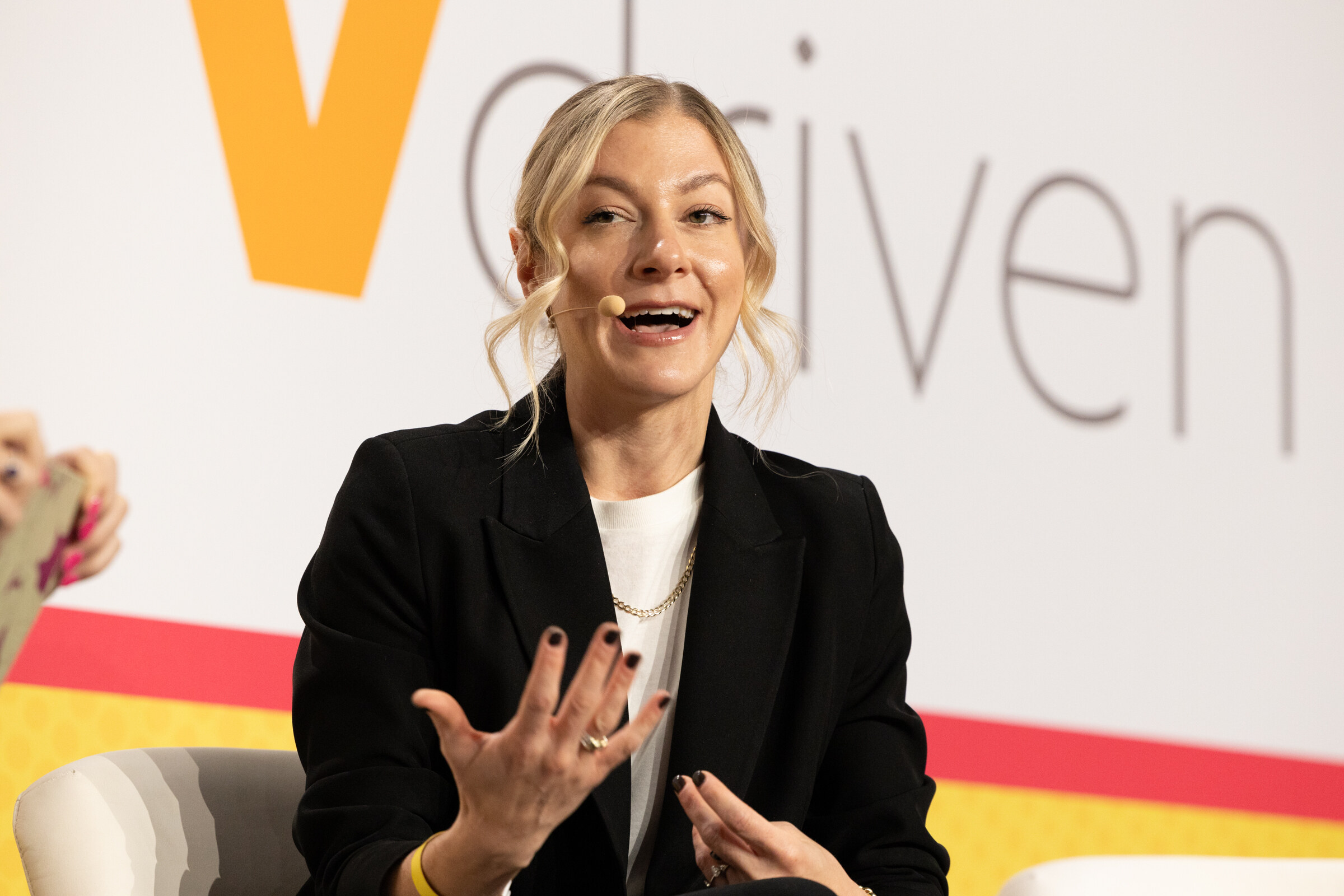 A blonde woman in a black blazer gesturing with both hands in front of a Vdriven backdrop.