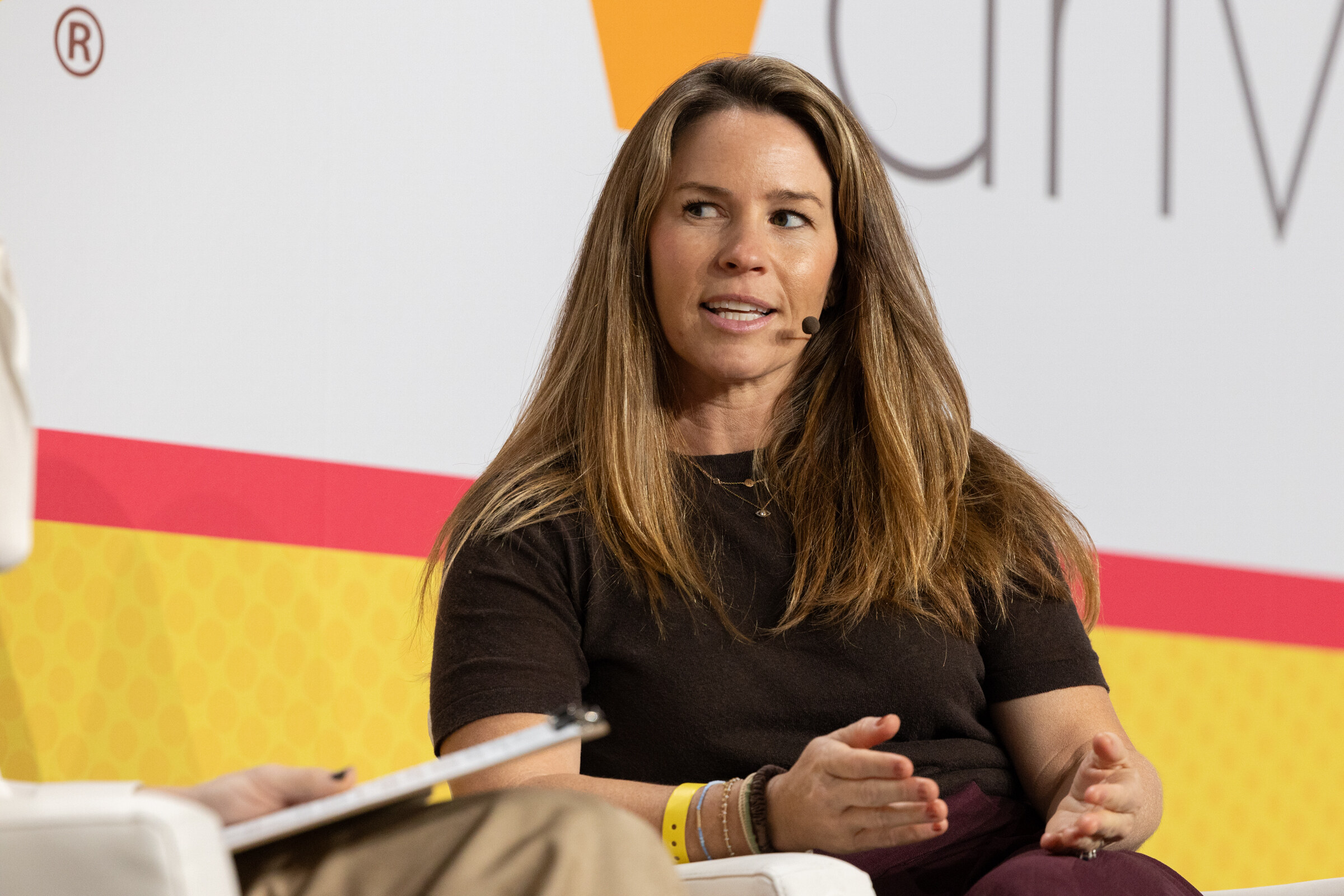 A woman with long brown hair in a dark top speaking on stage in front of a Vdriven backdrop.