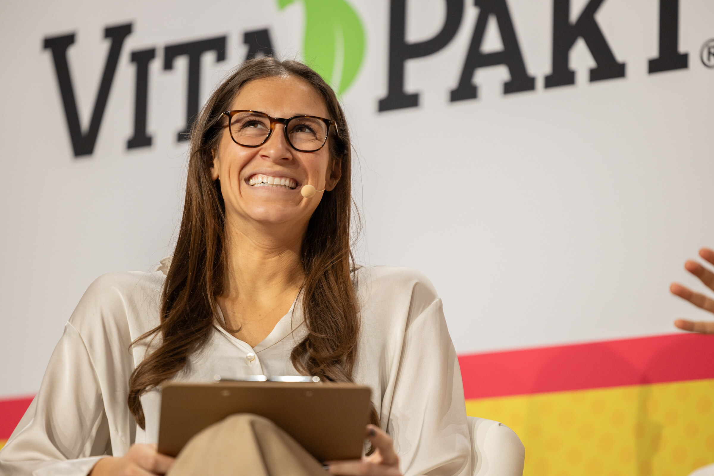 A smiling woman in glasses and a cream blouse holding a clipboard in front of a Vita-Pakt sign.