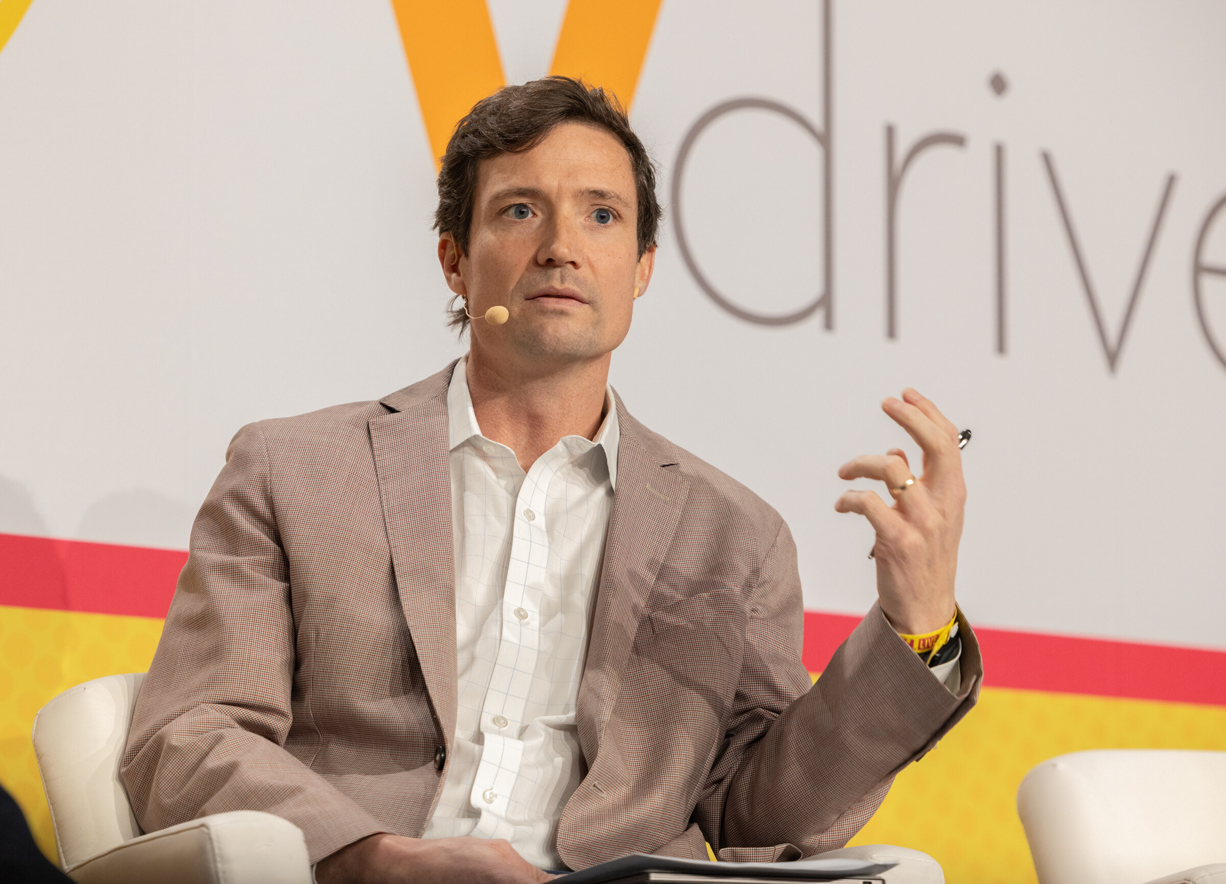 A man in a tan blazer holding a pen and gesturing in front of a Vdriven backdrop.