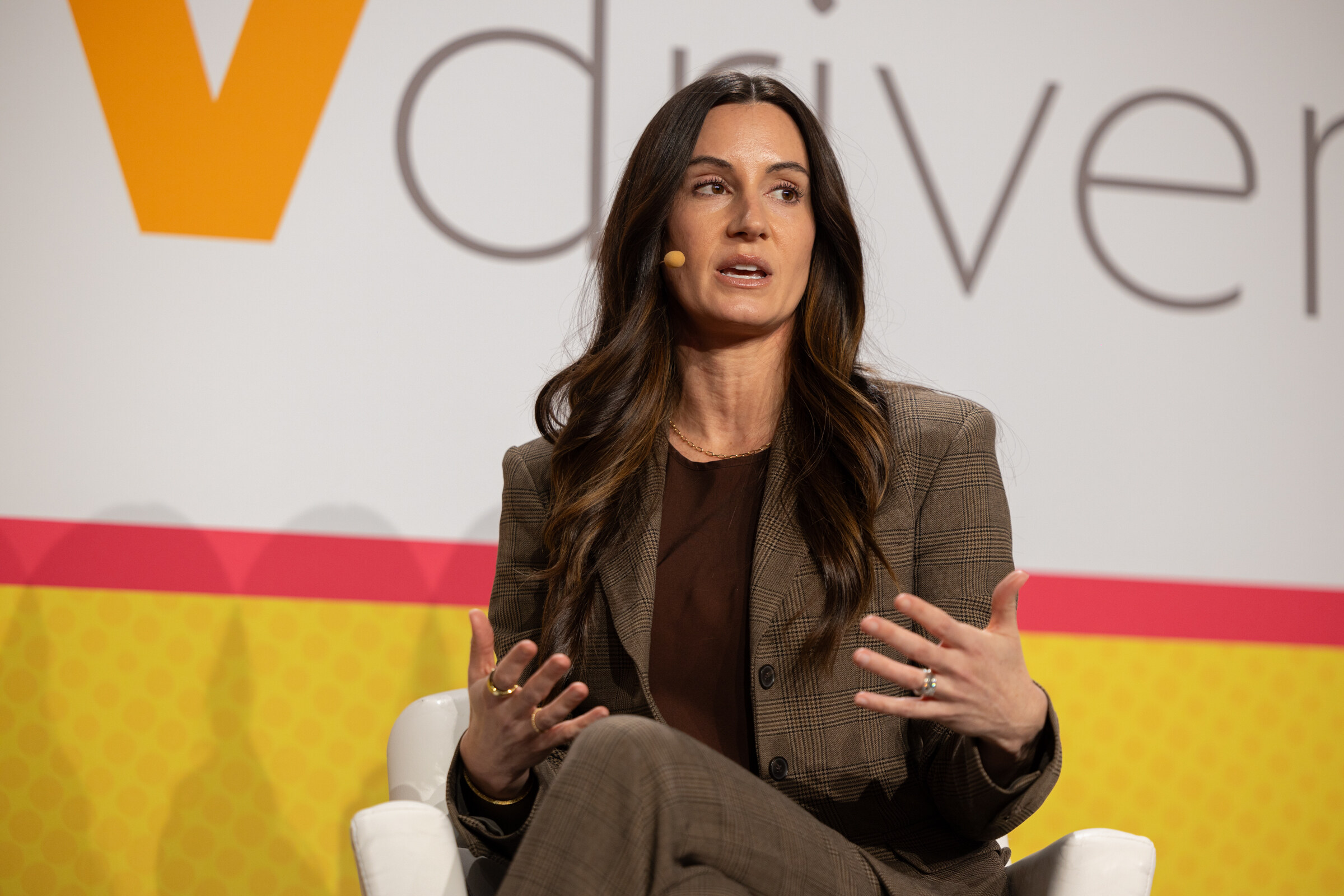 A woman with long dark hair in a brown blazer gesturing as she speaks in front of a Vdriven backdrop.