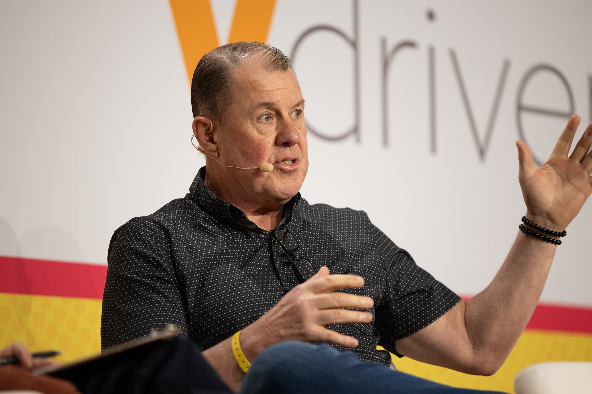 A man in a dark polo shirt raising one hand while speaking in front of a Vdriven backdrop.
