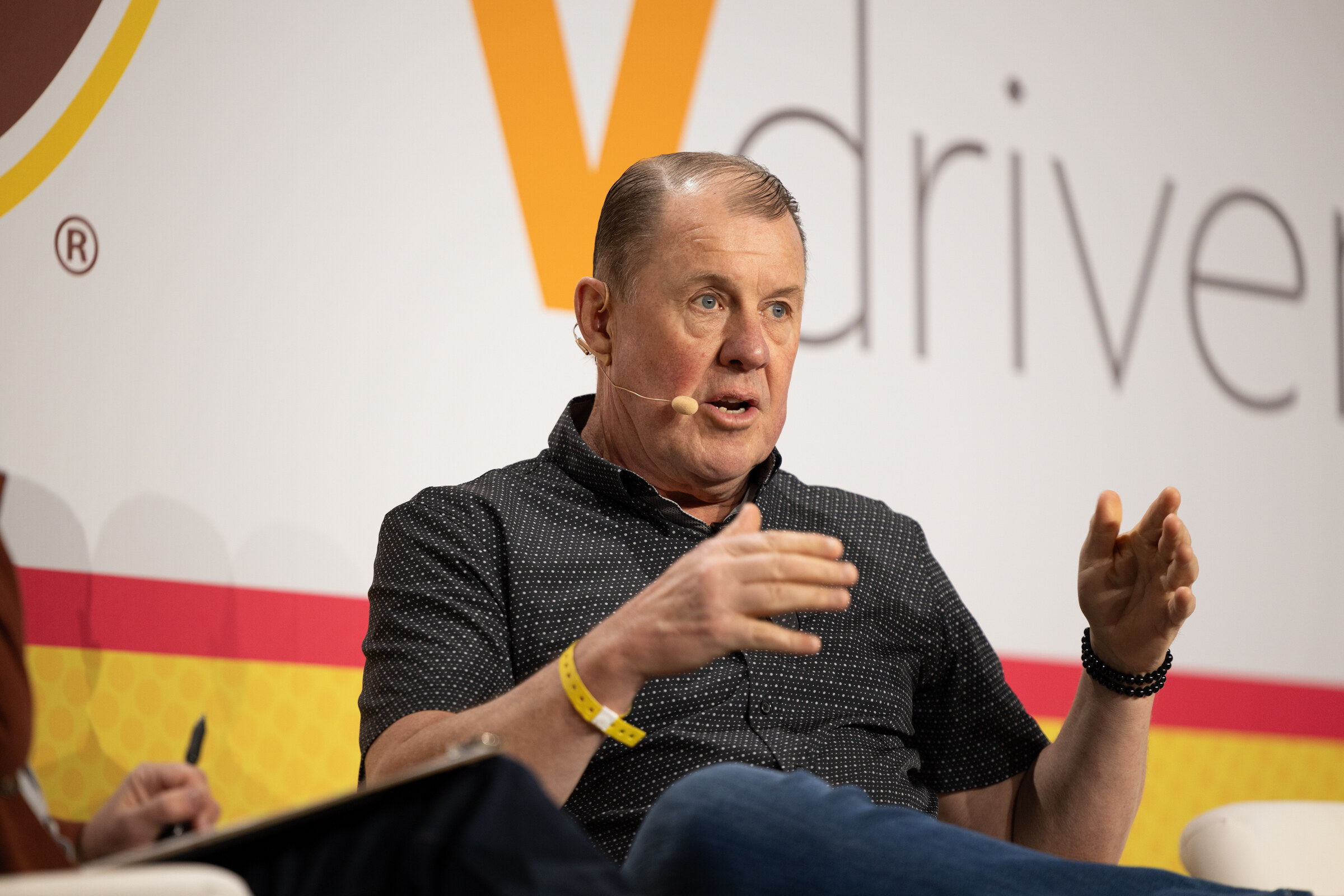 A man in a dark polo with beaded bracelets gesturing with both hands in front of a Vdriven backdrop.