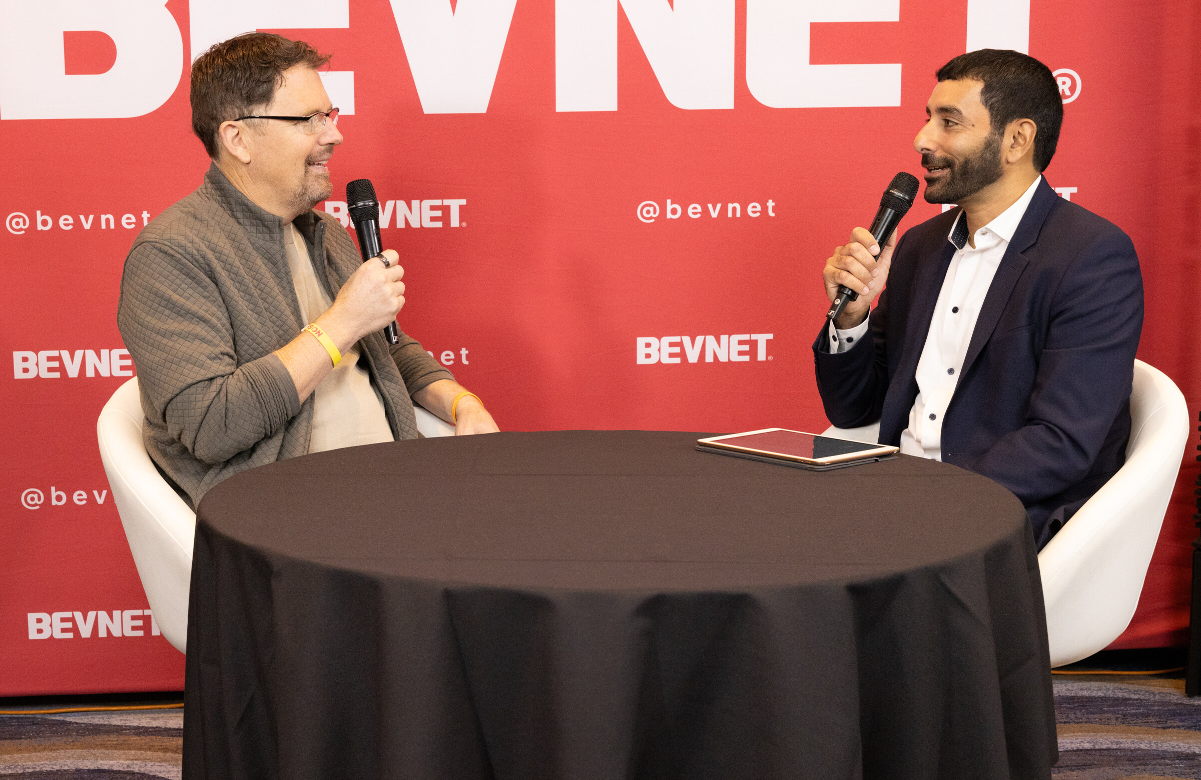 Two men with microphones seated at a small black table in front of a red BevNET backdrop.