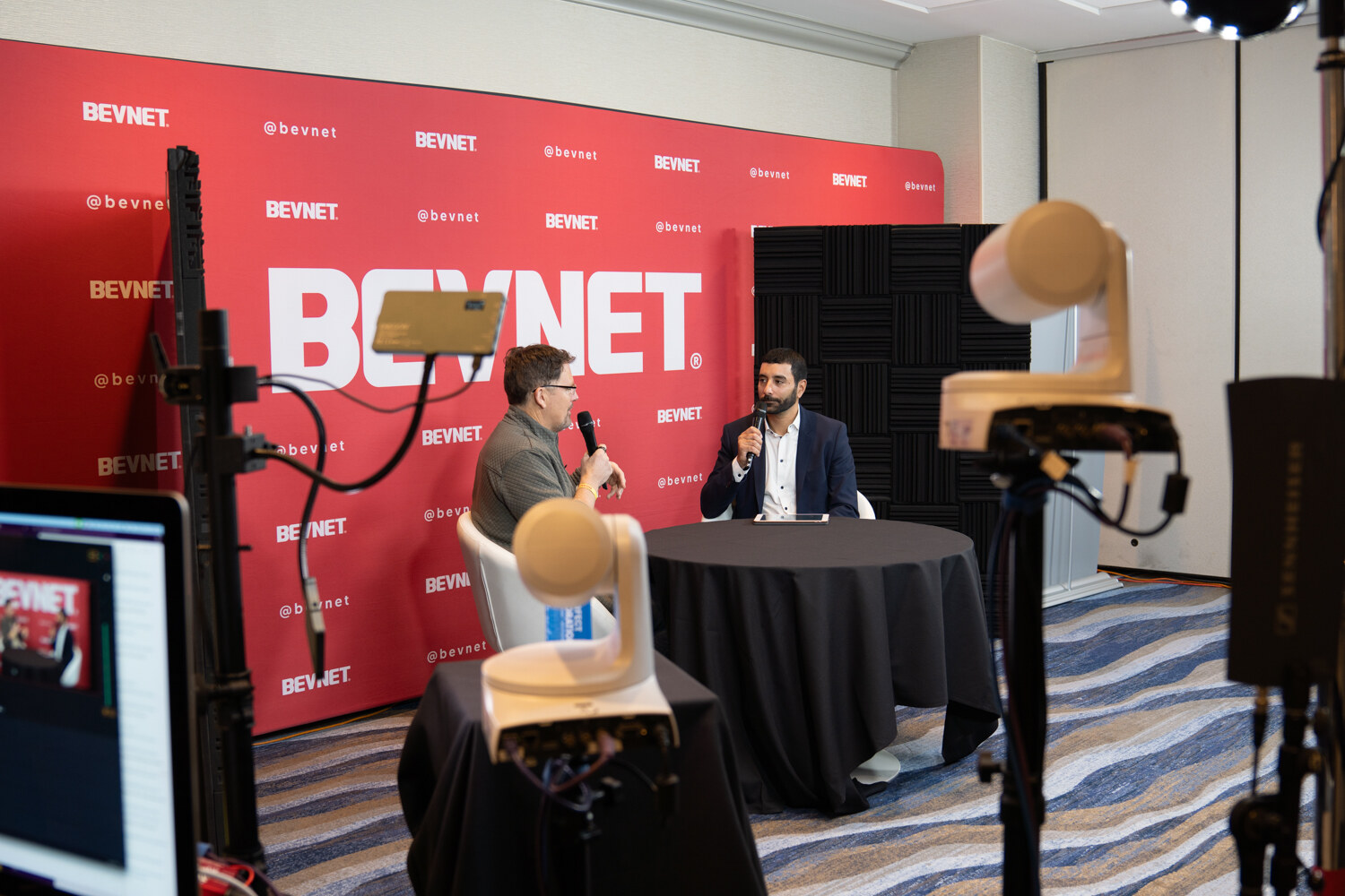 A red BevNET backdrop with two men in conversation at a small table, with two camera heads and a monitor in the foreground.