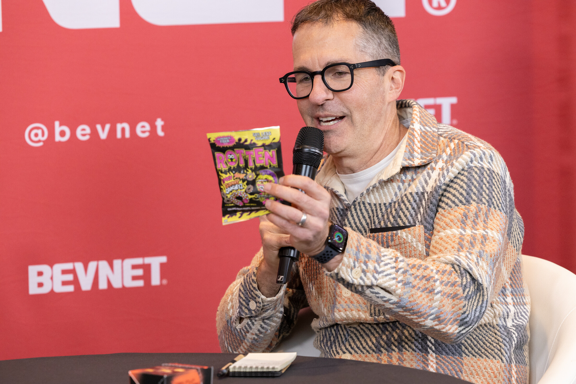 A man in a plaid jacket and black glasses holding a microphone and a bag of Rotten candy against a red BevNET backdrop.