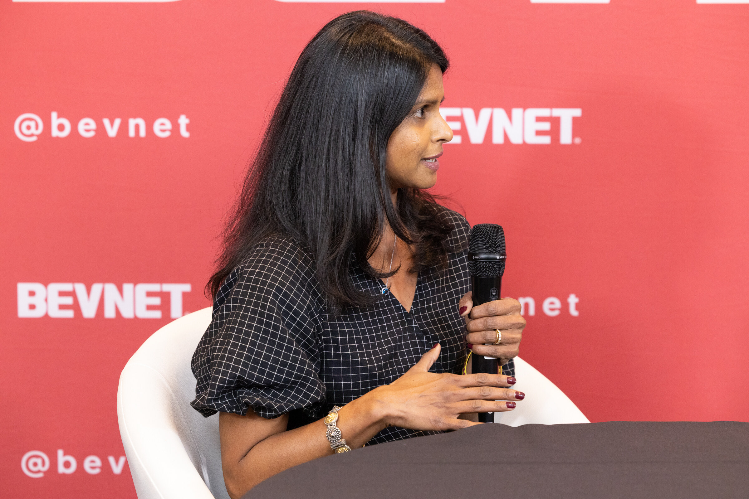 A woman with long dark hair in a checked top holding a microphone and speaking in front of a BevNET backdrop.
