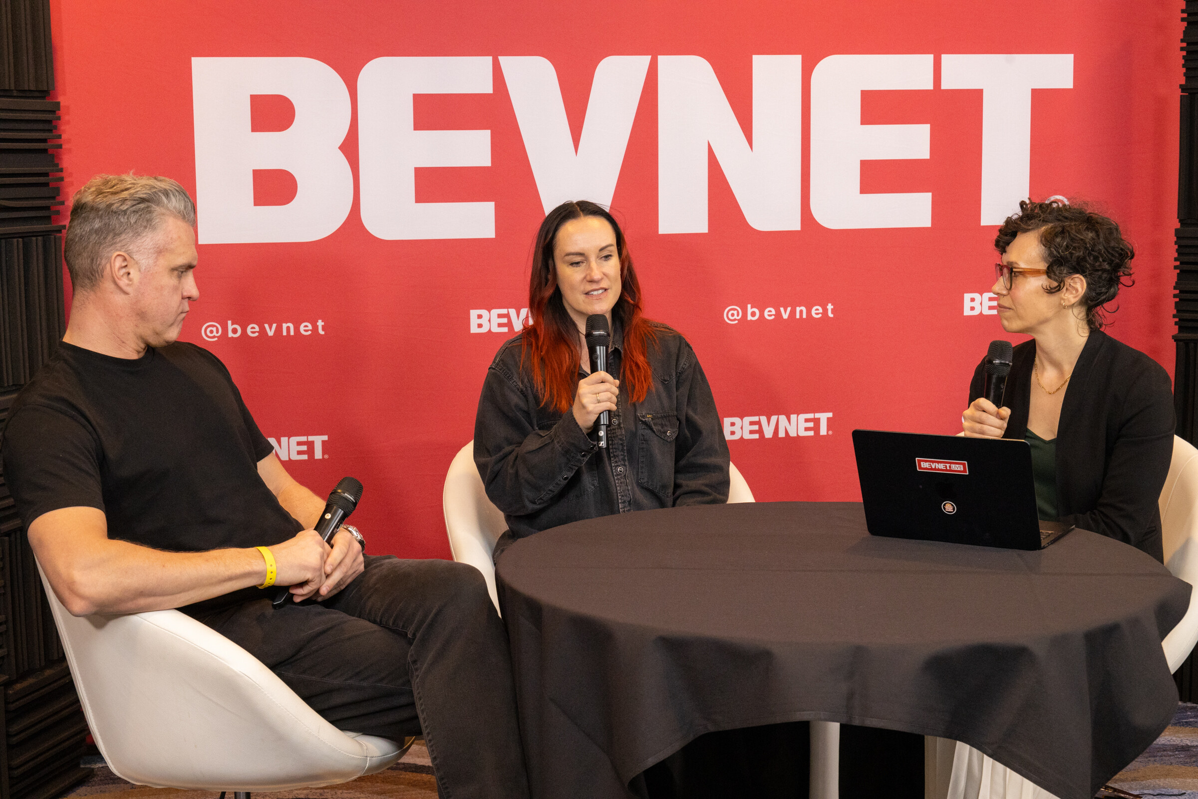 A man in a black t-shirt, a woman with a microphone and a woman with a laptop around a small table in front of a BevNET backdrop.