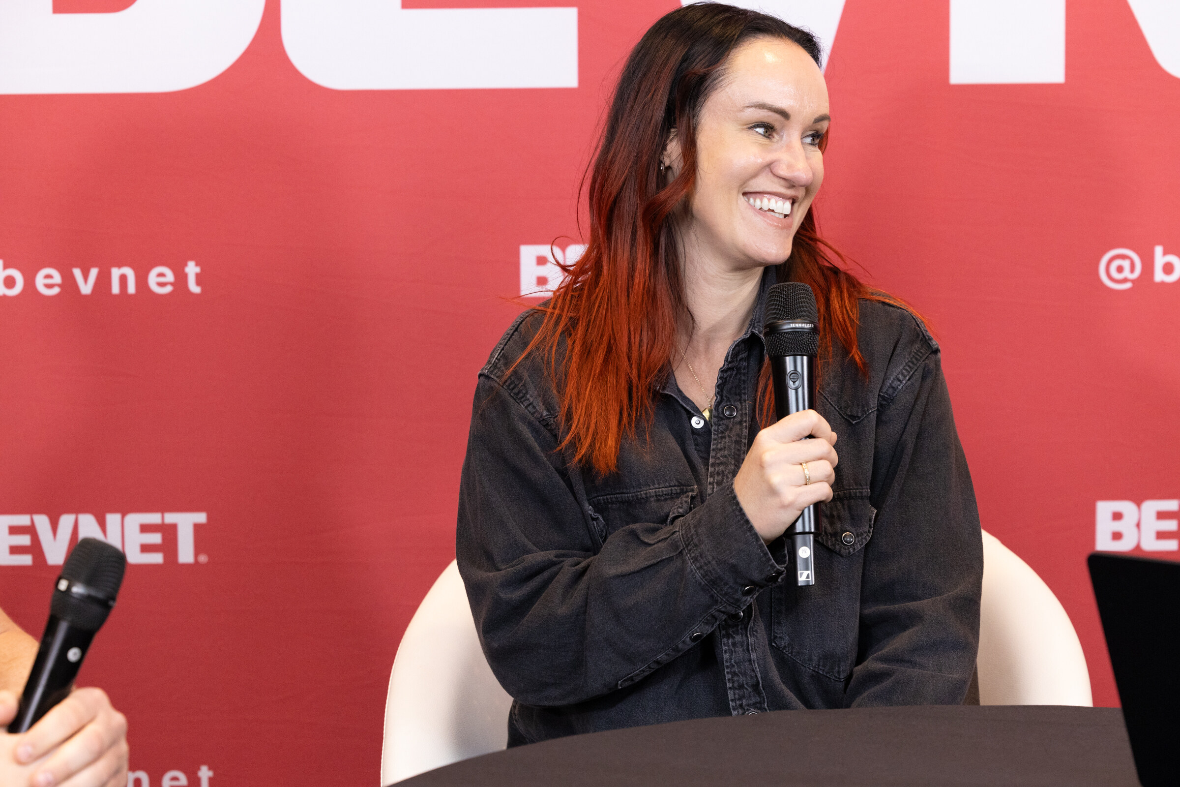 A smiling woman with red-brown hair in a dark denim jacket holding a microphone in front of a BevNET backdrop.
