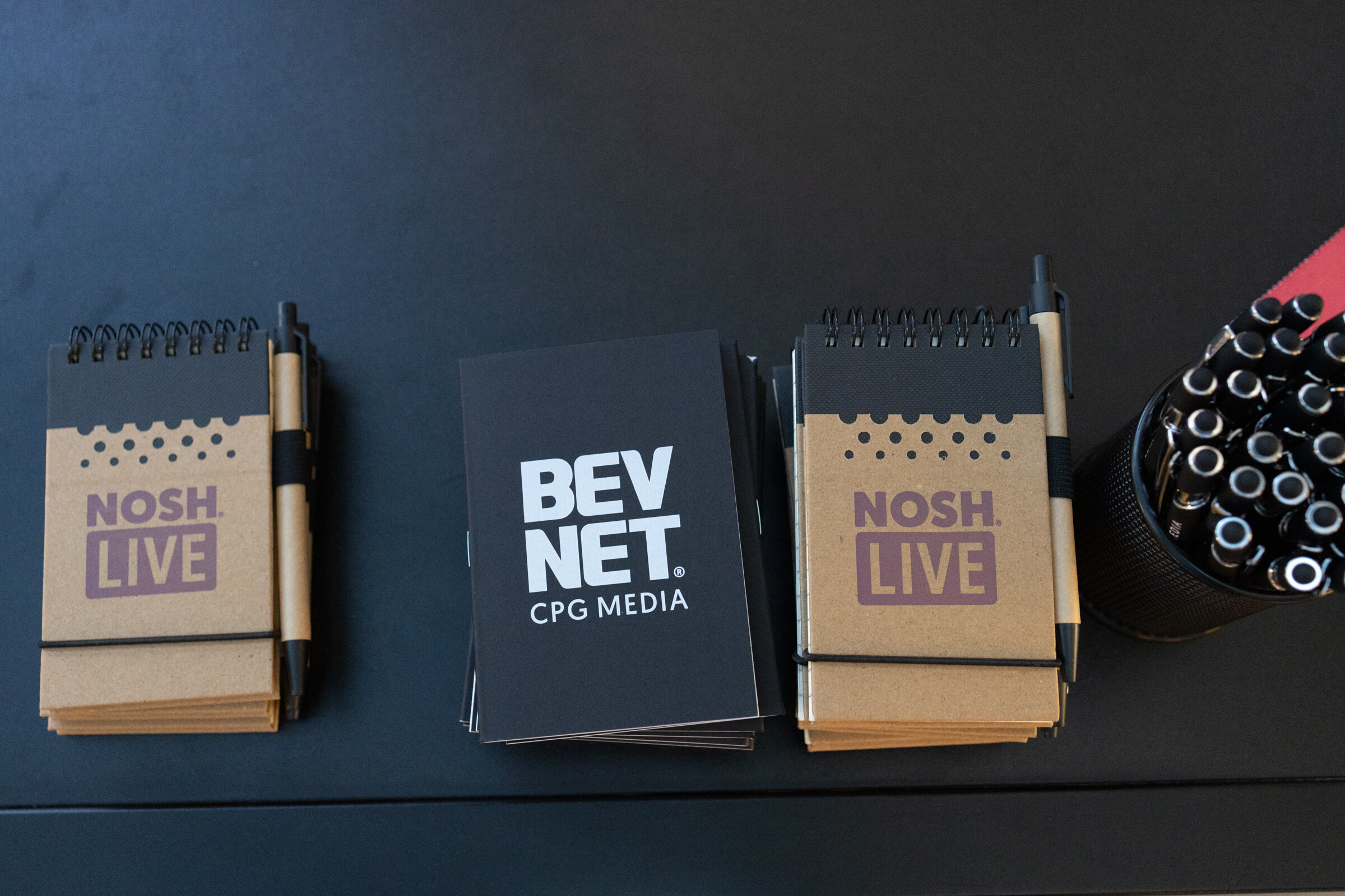 Notebooks marked Nosh Live and BevNET CPG Media with pens on a dark table.
