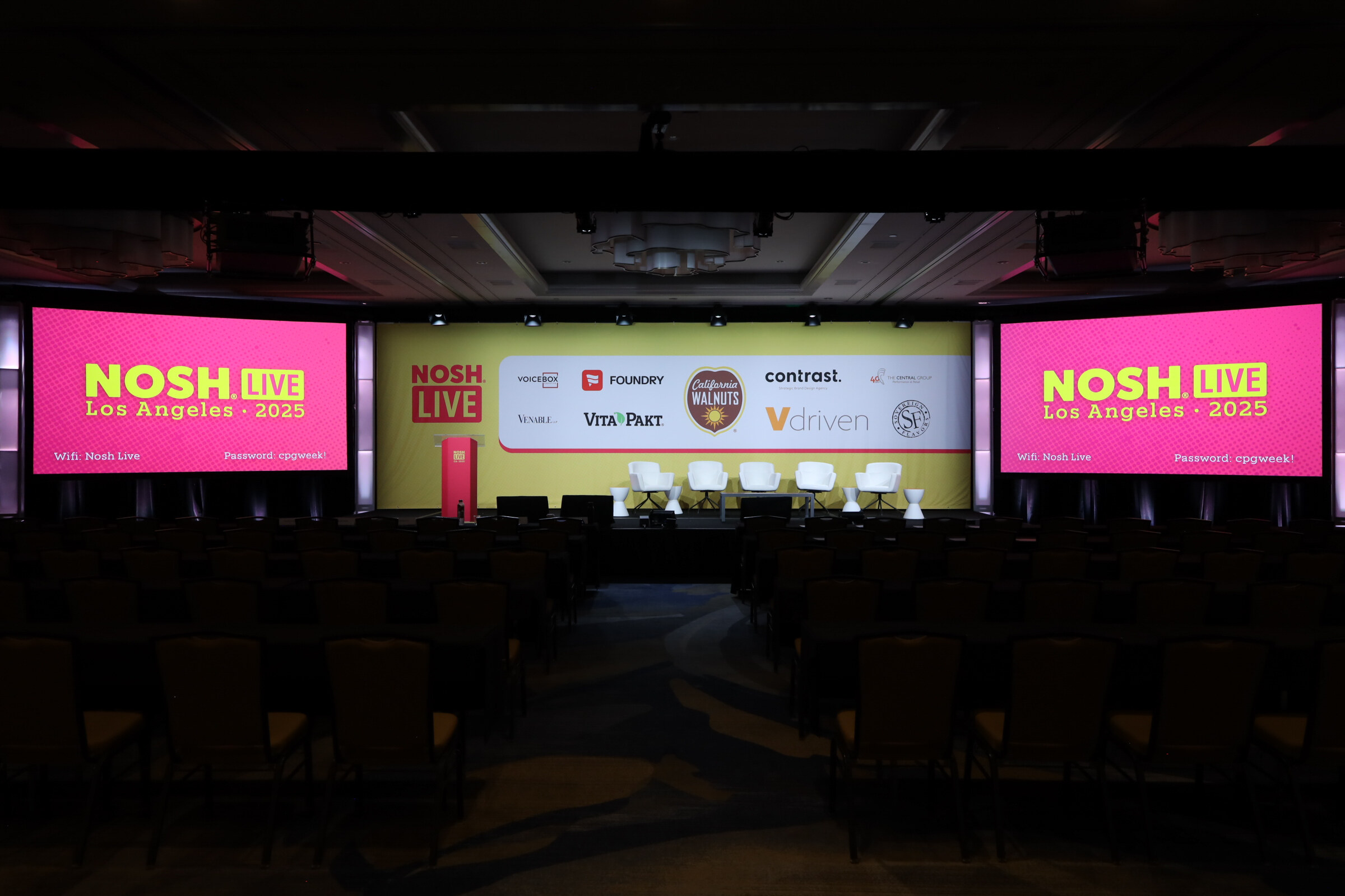 A darkened ballroom with a stage backdrop of sponsor logos and two pink screens reading Nosh Live Los Angeles 2025.